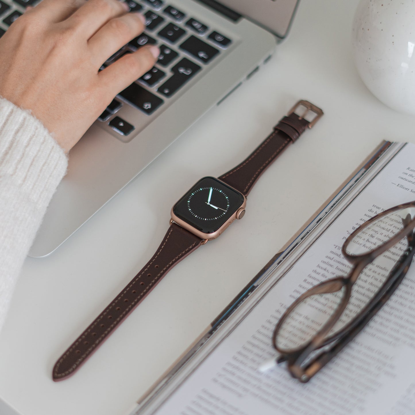 Eine Person tippt auf einem Laptop neben einem Arktisband Apple Watch Lederarmband „Napoli“, einer offenen Zeitschrift, einer Brille und einer weißen Tasse – alles minimalistisch auf einem weißen Schreibtisch angeordnet.