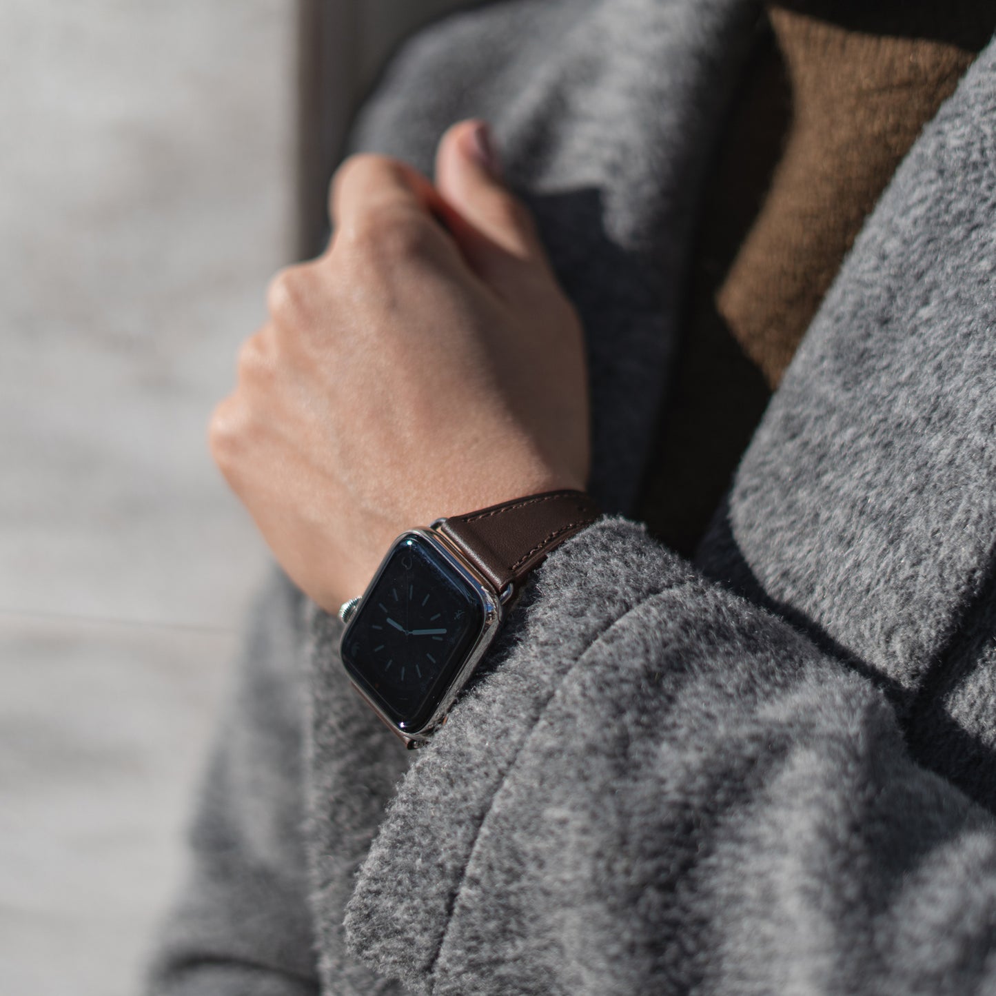 Eine Person in grauem Mantel und braunem Oberteil, von den Schultern abwärts gesehen, checkt die Uhrzeit auf einem Arktisband Apple Watch Lederarmband „Napoli“. Der Hintergrund ist unscharf.