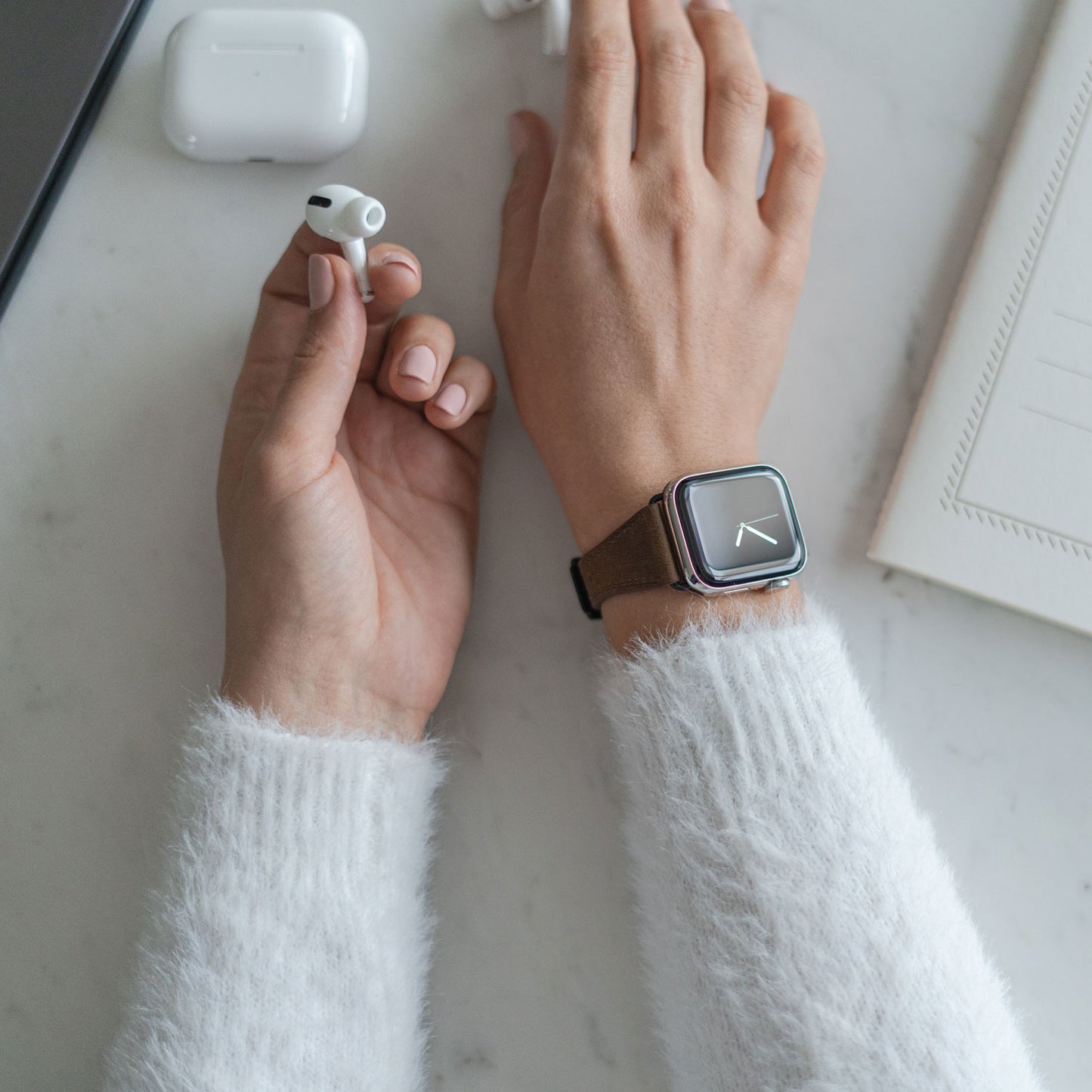 Eine Person in einem weißen Flauschpullover und einem Arktisband Apple Watch Lederarmband „Napoli“ hält einen kabellosen Ohrhörer. Auf der weißen Fläche daneben liegen ein geöffnetes Ladecase und ein Notebook.