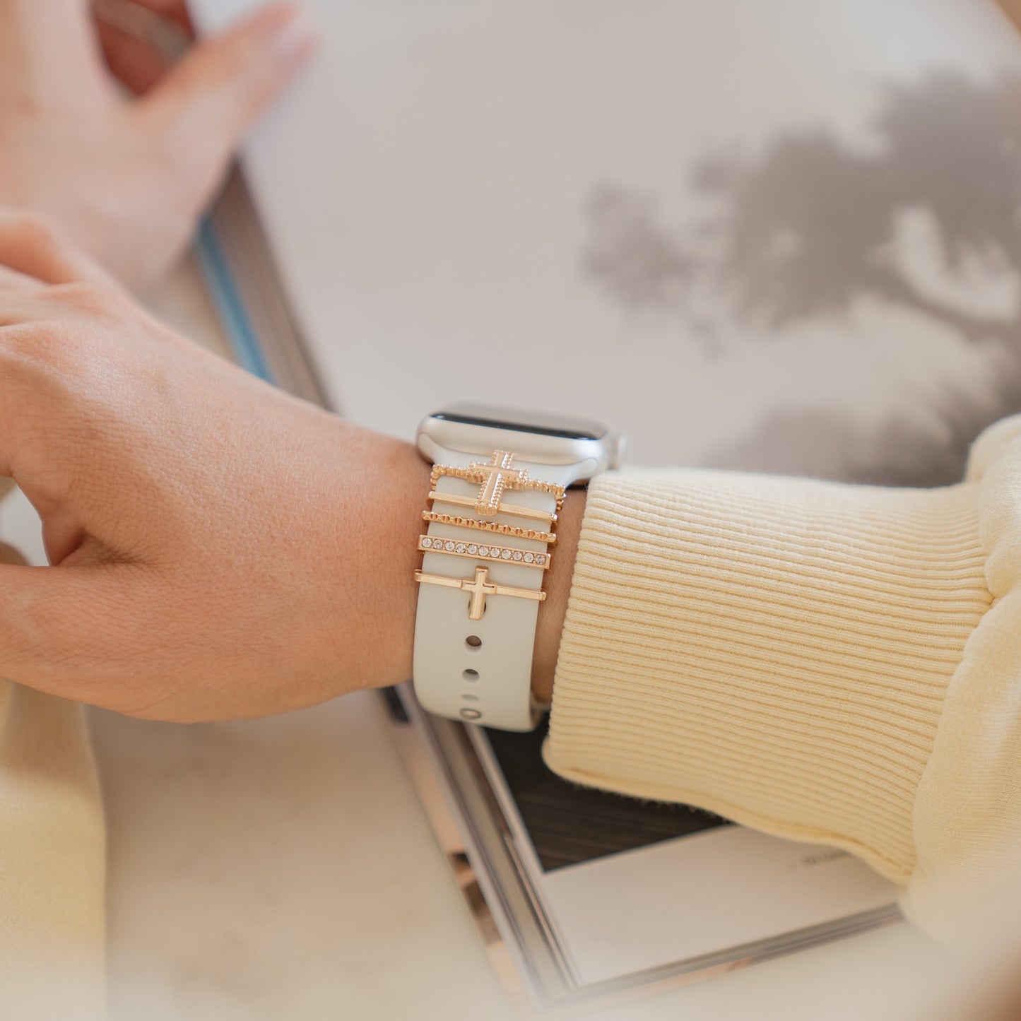 Jemand trägt ein leichtes Langarmshirt und kombiniert seine Apple Watch mit einem weißen Arktisband-Armband und mehreren goldenen und juwelenbesetzten Arktisband-Apple-Watch-Armbandanhängern. Im Hintergrund ist ein verschwommenes Buch oder eine Zeitschrift zu sehen.