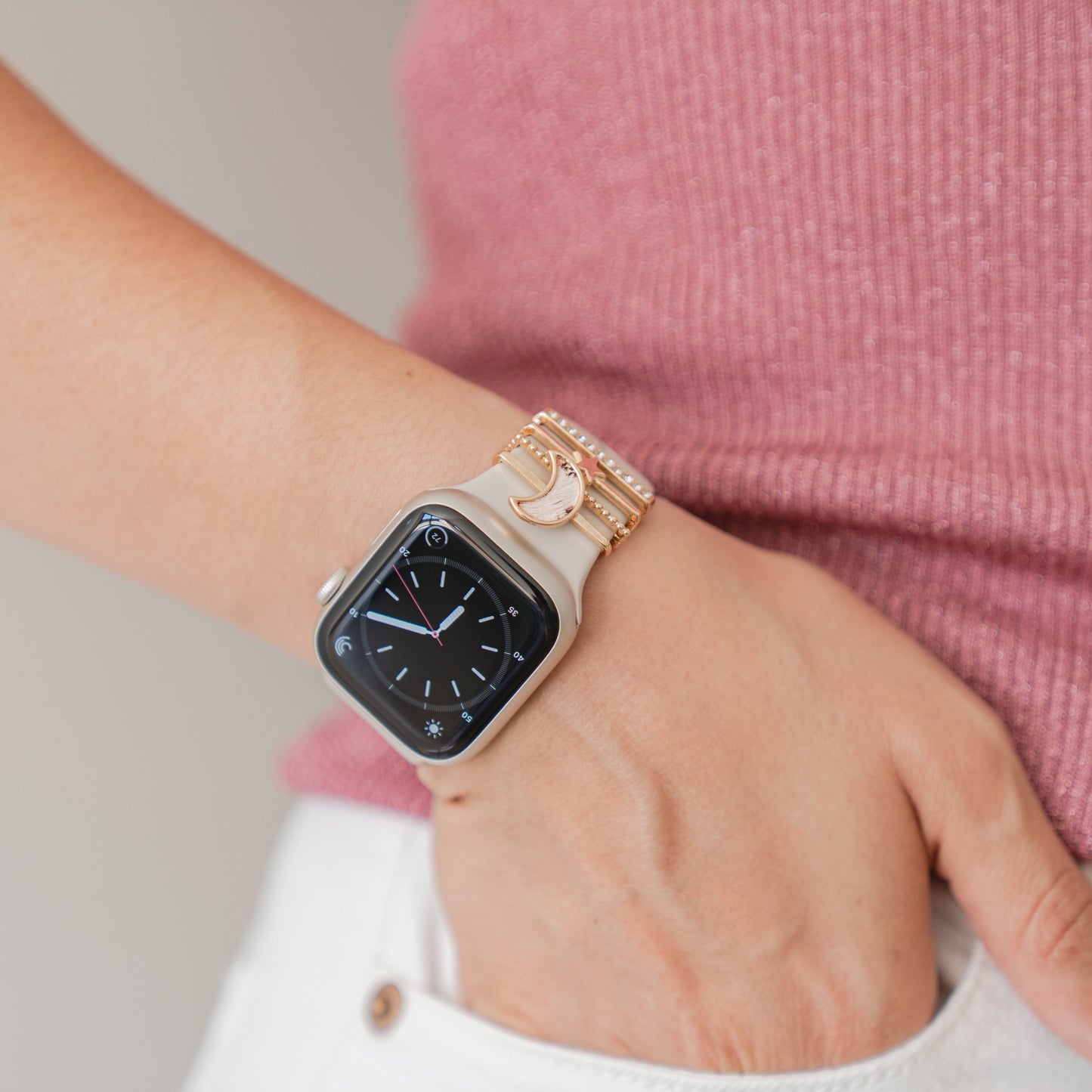 Eine Person trägt ein Arktisband Apple Watch Armband Charms und ein goldenes Armband mit einem Halbmond-Anhänger am linken Handgelenk und ist mit einem rosa gerippten Hemd und weißen Hosen bekleidet.