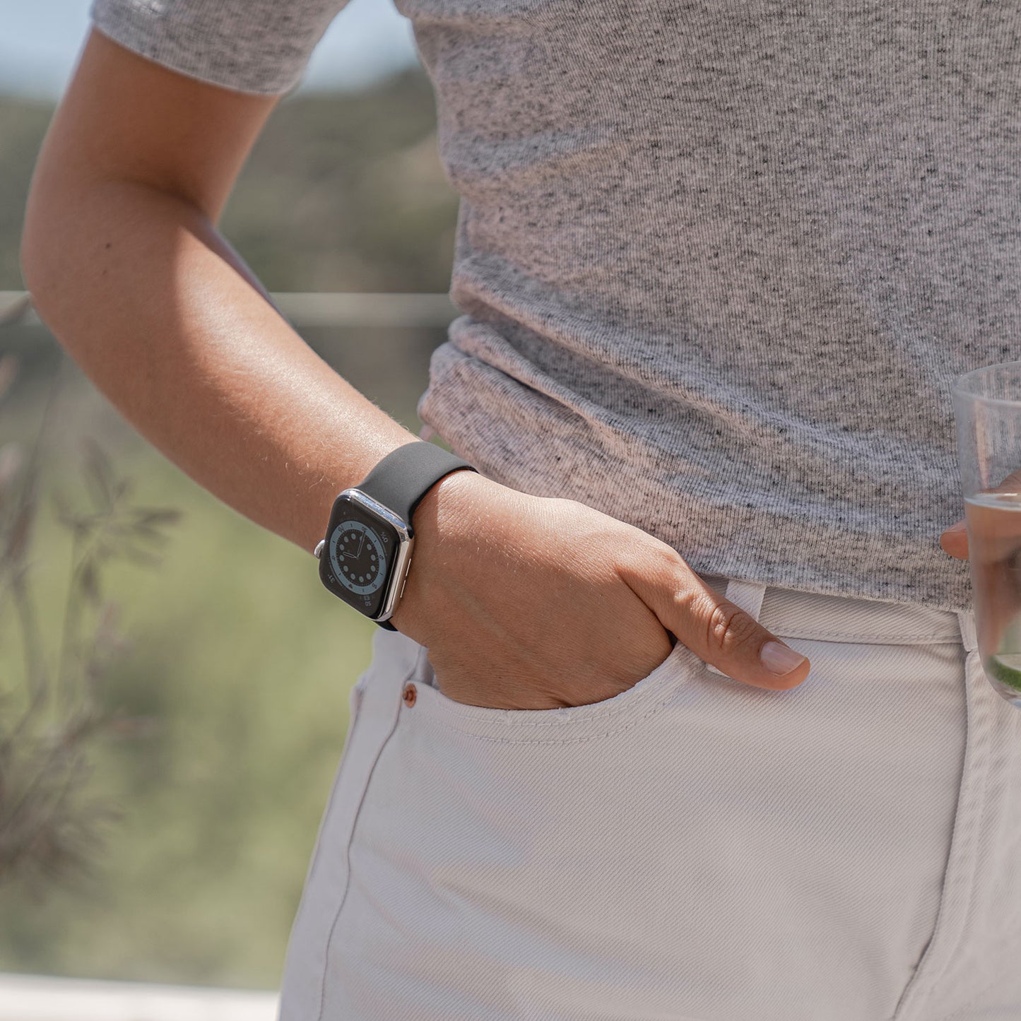 Eine Person in einem grauen Kurzarmhemd und einer hellen Hose steht mit einer Hand in der Tasche, trägt das arktisband Apple Watch Silikonarmband „Aqua“ und hält in der anderen Hand ein Glas mit einem Getränk.