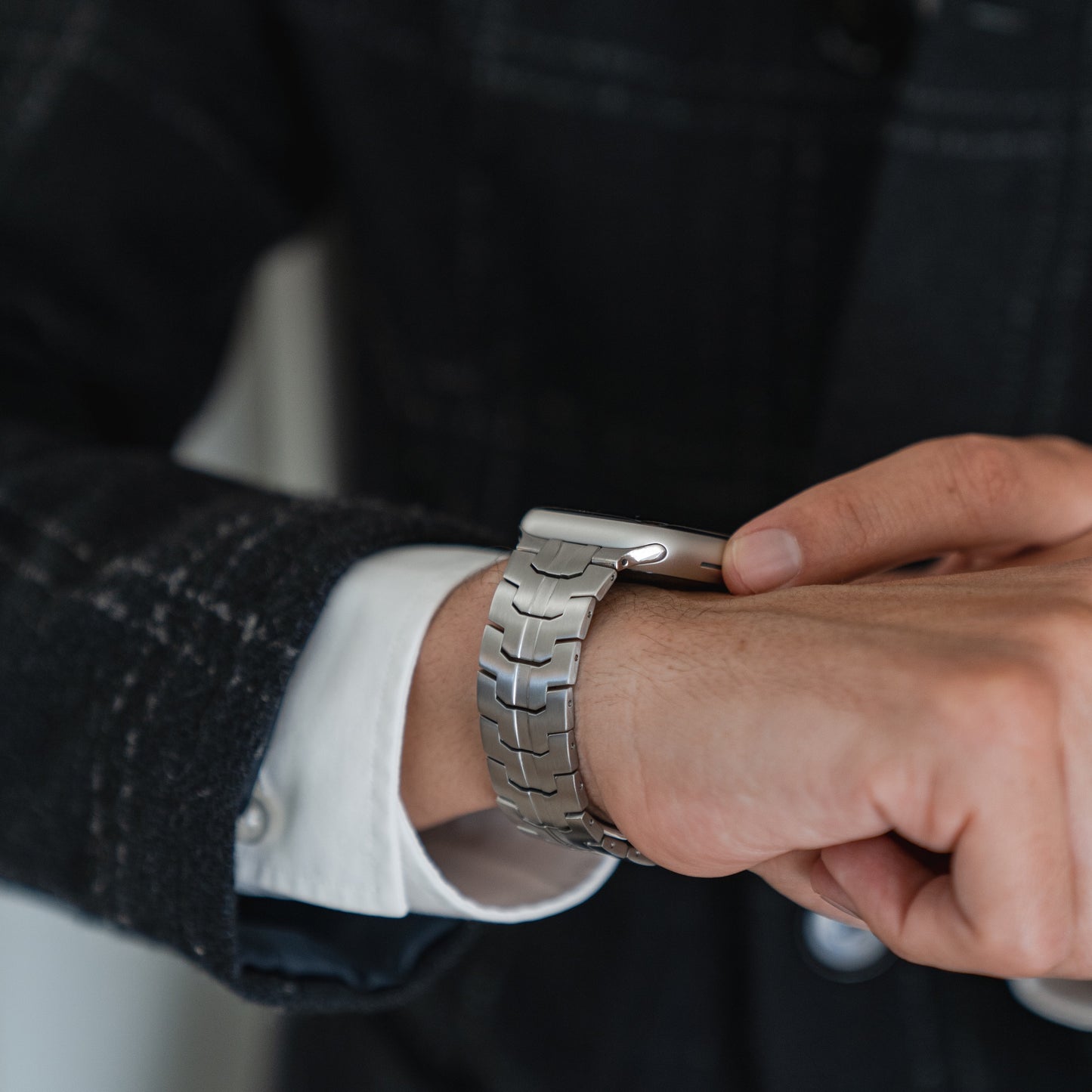 Eine Person in einer dunkelkarierten Jacke und einem weißen Hemd rückt ihr Arktisband Apple Watch Edelstahlarmband „Momento“ an ihrem linken Handgelenk zurecht und berührt dabei mit der rechten Hand das silberne Band.