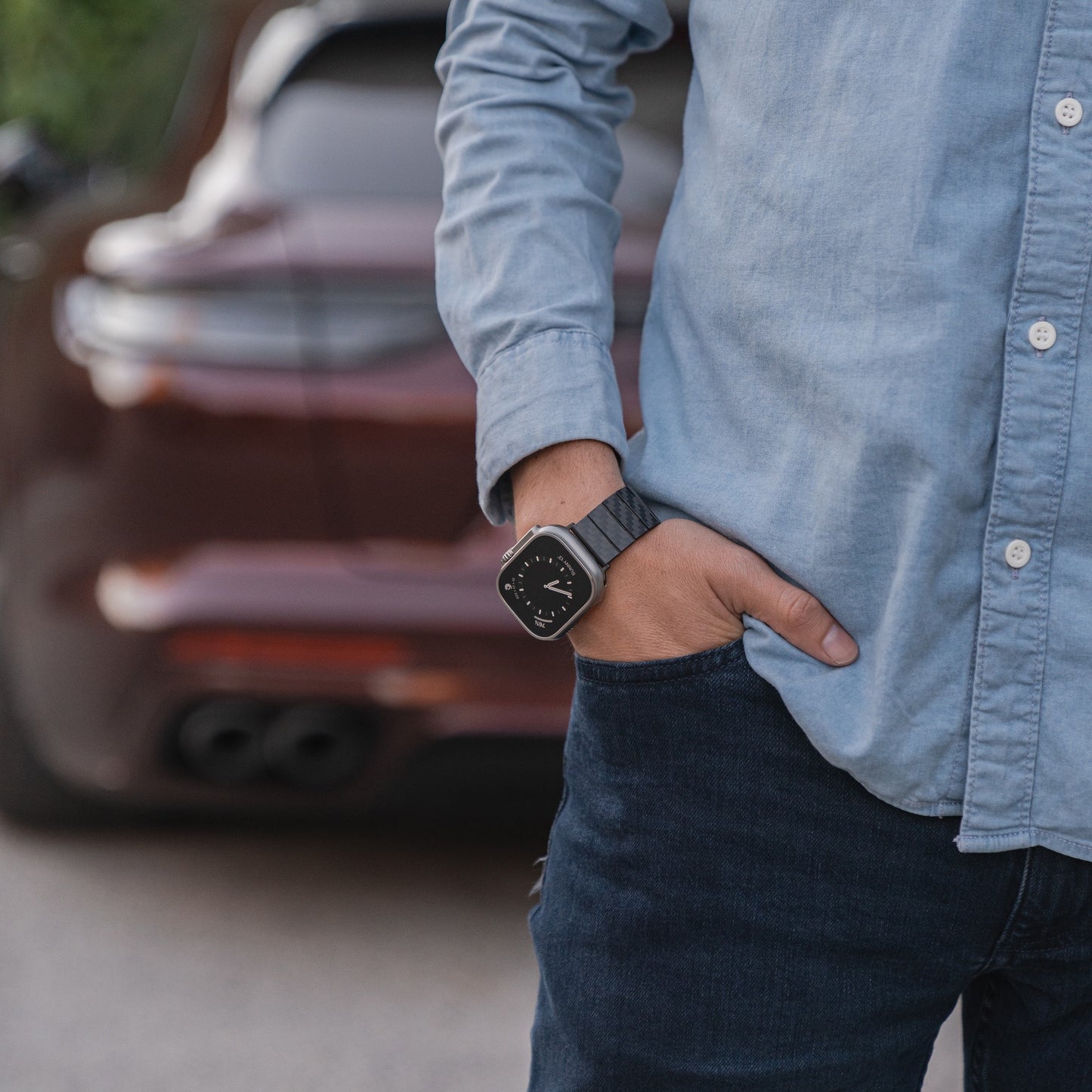 Eine Person in blauem Hemd und Jeans steht mit der Hand in der Tasche und trägt das arktisband Apple Watch Ultra KARBON Armband aus echter Karbonfaser, nahe einem geparkten Auto.