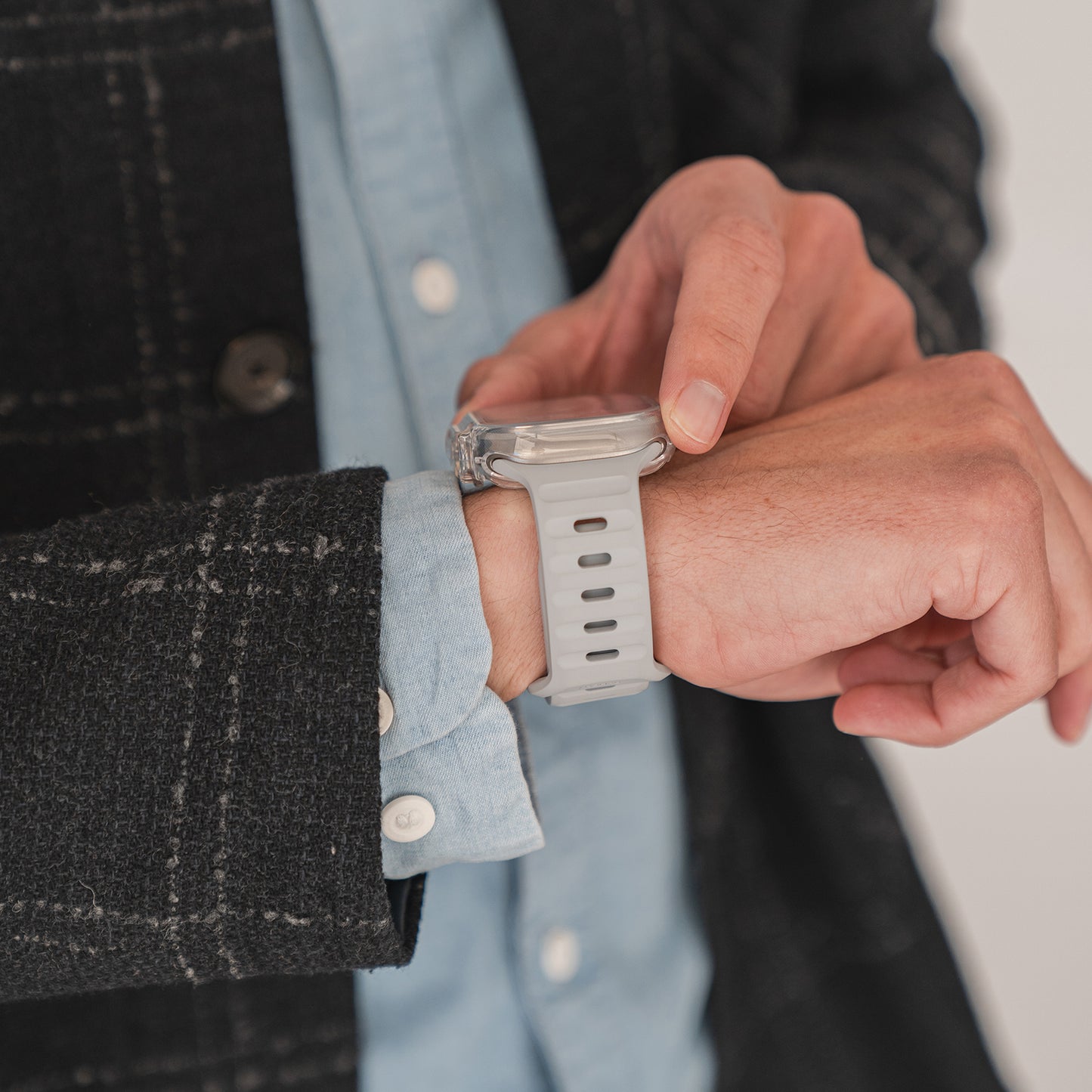 Eine Person, die das arktis.de ArktisPRO Invisible Air Case für die Apple Watch Ultra mit weißem Armband trägt, das einen 360° flexiblen TPU-Schutz bietet, berührt den Bildschirm, während sie eine schwarze Jacke und ein blaues Hemd trägt.