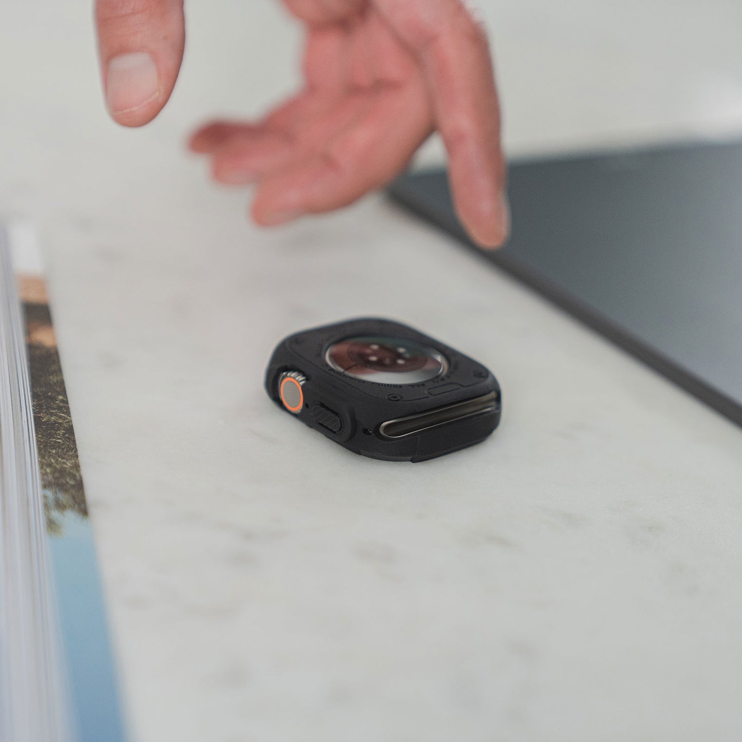 Eine Hand greift nach einer kleinen schwarzen Action-Kamera auf einer weißen Oberfläche in der Nähe eines Laptops und einer Zeitschrift mit dem ArktisPRO SHOCKPROOF Case für die Apple Watch Ultra 49 mm von ArktisPRO.