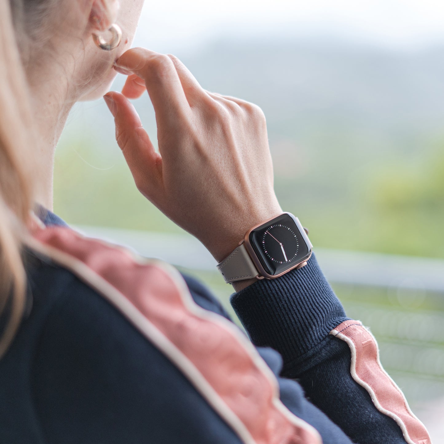 Eine Person im Freien, gekleidet in eine marineblaue und rosa Jacke, trägt das arktisband Apple Watch Lederarmband „Napoli“ mit beigem Band und dunklem, minimalistischem Zifferblatt und berührt nachdenklich ihr Kinn.