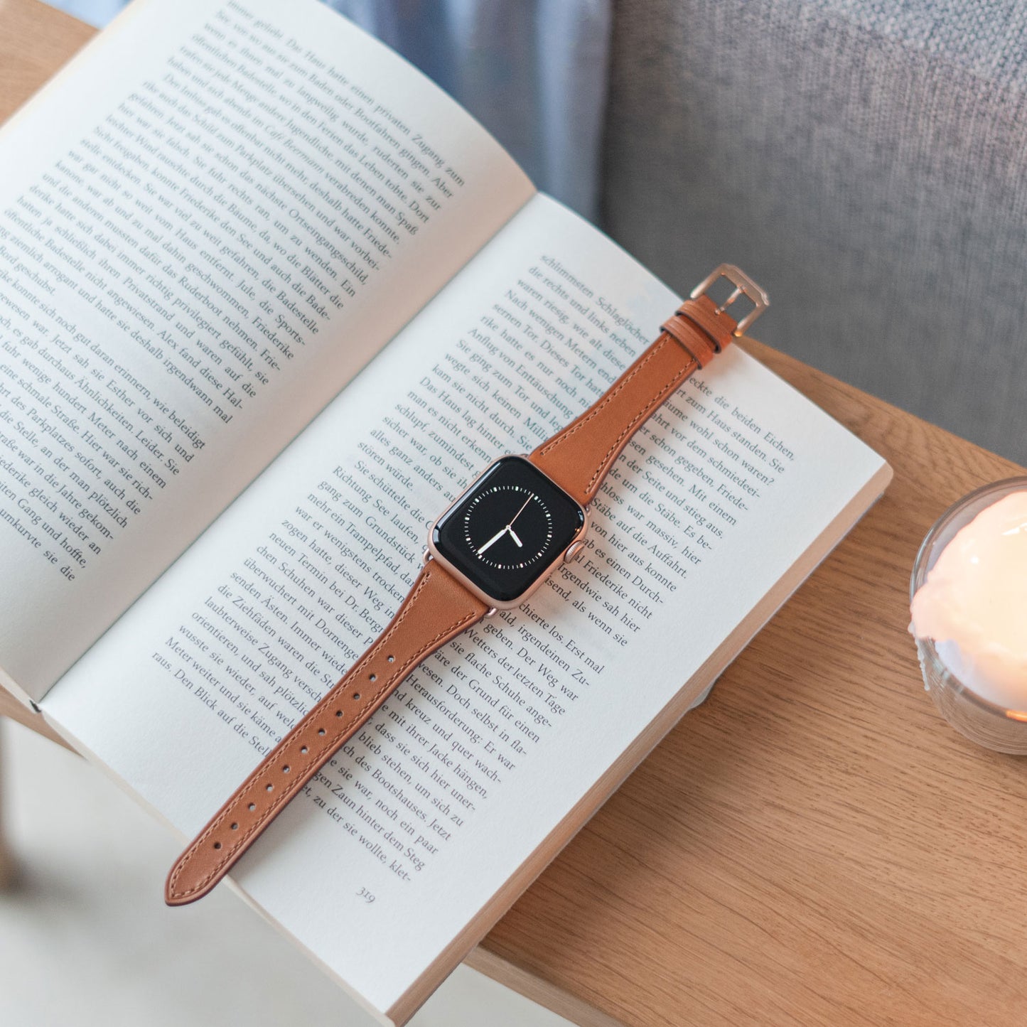 Das arktisband Apple Watch Lederarmband „Napoli“ von arktisband liegt im minimalistischen Stil auf einem Buch, neben einer brennenden Kerze auf einem Holztisch.