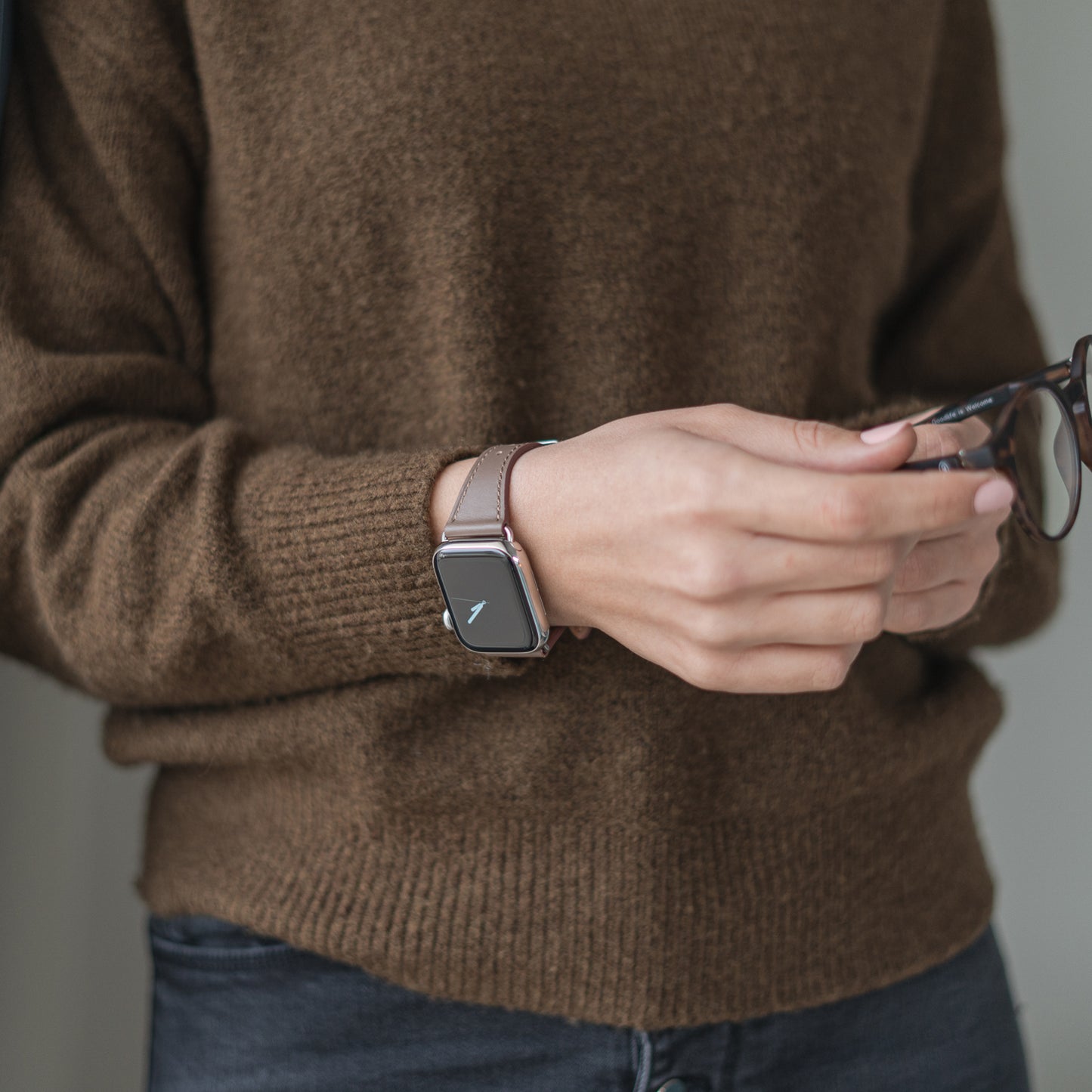 Eine Person in einem braunen Pullover und dunkler Jeans hält in der einen Hand eine Brille und trägt am anderen Handgelenk das im minimalistischen Stil gehaltene arktisband Apple Watch Lederarmband „Napoli“ von arktisband.