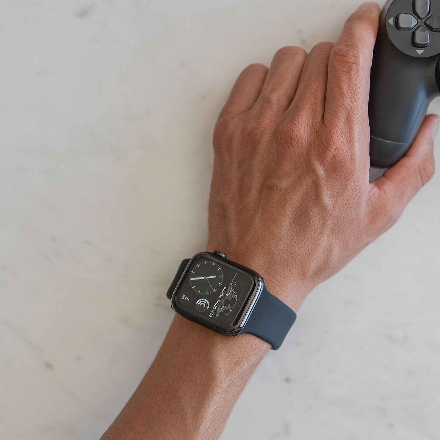 Jemand trägt ein arktisband Apple Watch Silikonarmband „Aqua“ von arktisband am linken Handgelenk und hält mit der rechten Hand einen schwarzen Videospiel-Controller auf einer hellen Marmoroberfläche.