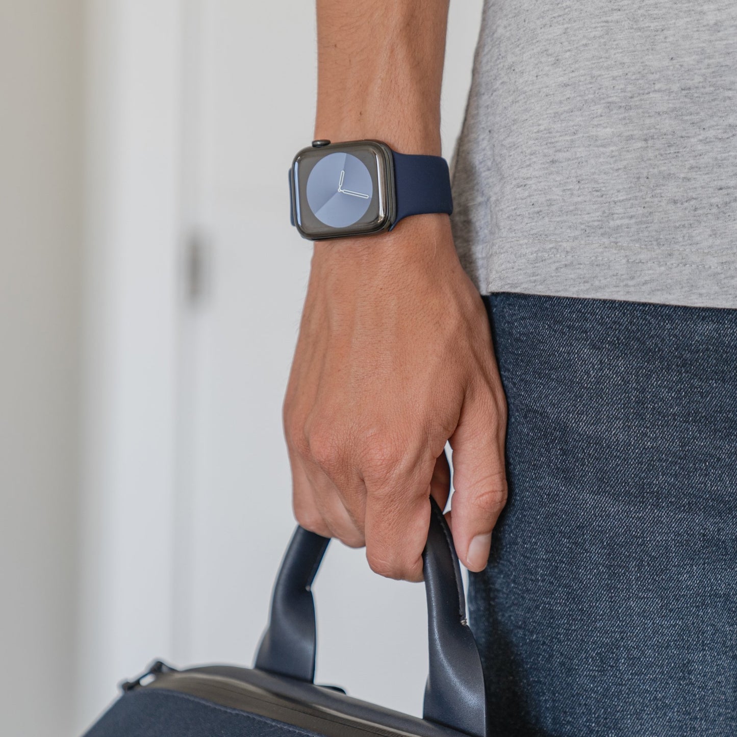 Eine Person in grauem Kurzarmhemd und dunkler Jeans hält eine dunkelblaue Tasche am Henkel. Sie trägt ein arktisband Apple Watch Silikonarmband „Aqua“ von arktisband, was ihrem Look eine sportliche Note verleiht.
