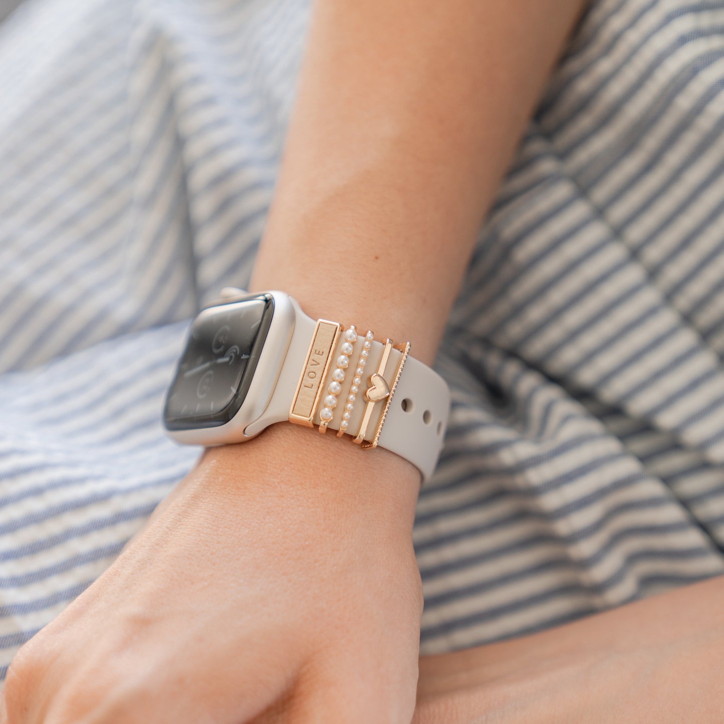 Eine Person trägt eine weiße Smartwatch mit dekorativen goldenen und perlenbesetzten Armbändern, die mit Arktisband Apple Watch Armband Charms von Arktisband verziert sind, in blauen und weißen Streifen gehalten sind und bei der das Uhrendisplay ausgeschaltet oder abgedunkelt ist.