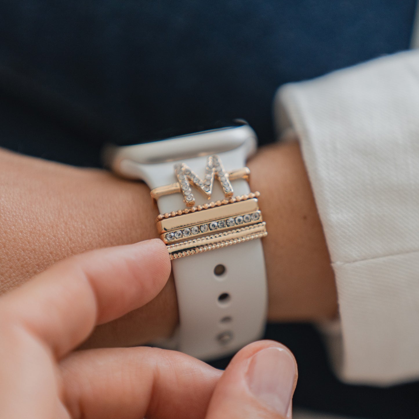 Nahaufnahme einer Hand, die eine Apple Watch mit Arktisband Apple Watch Armband-Anhängern einstellt. Diese bestehen aus einem goldenen Ring mit dem Buchstaben M und einem weiteren Ring mit kleinen klaren Steinen. Marke: Arktisband.