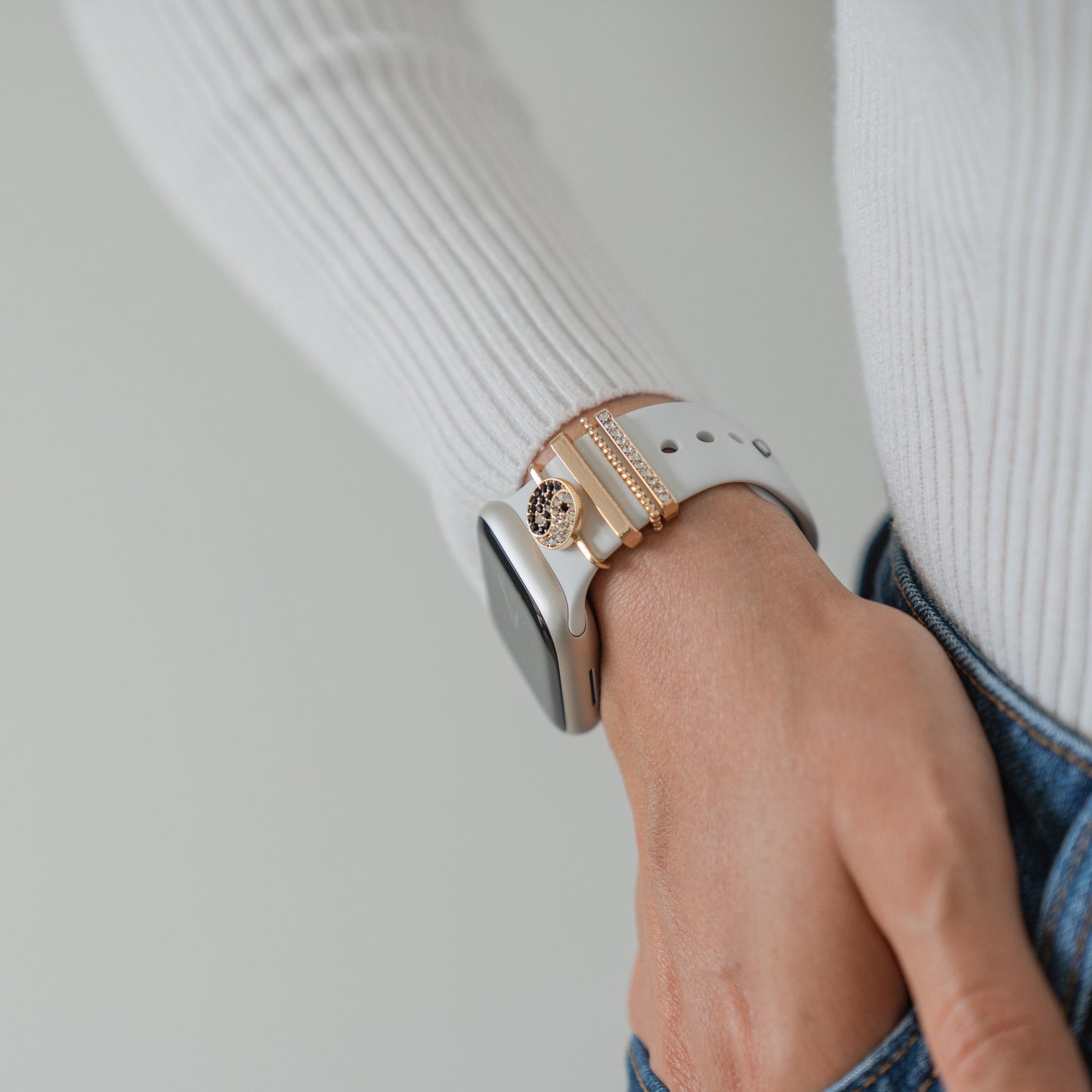 Eine Person in einem weißen Langarmshirt und Bluejeans, die Hand in der Tasche, trägt als Accessoires Arktisband-Apple-Watch-Armbandanhänger, eine weiße Smartwatch und mehrere goldene Armbänder.
