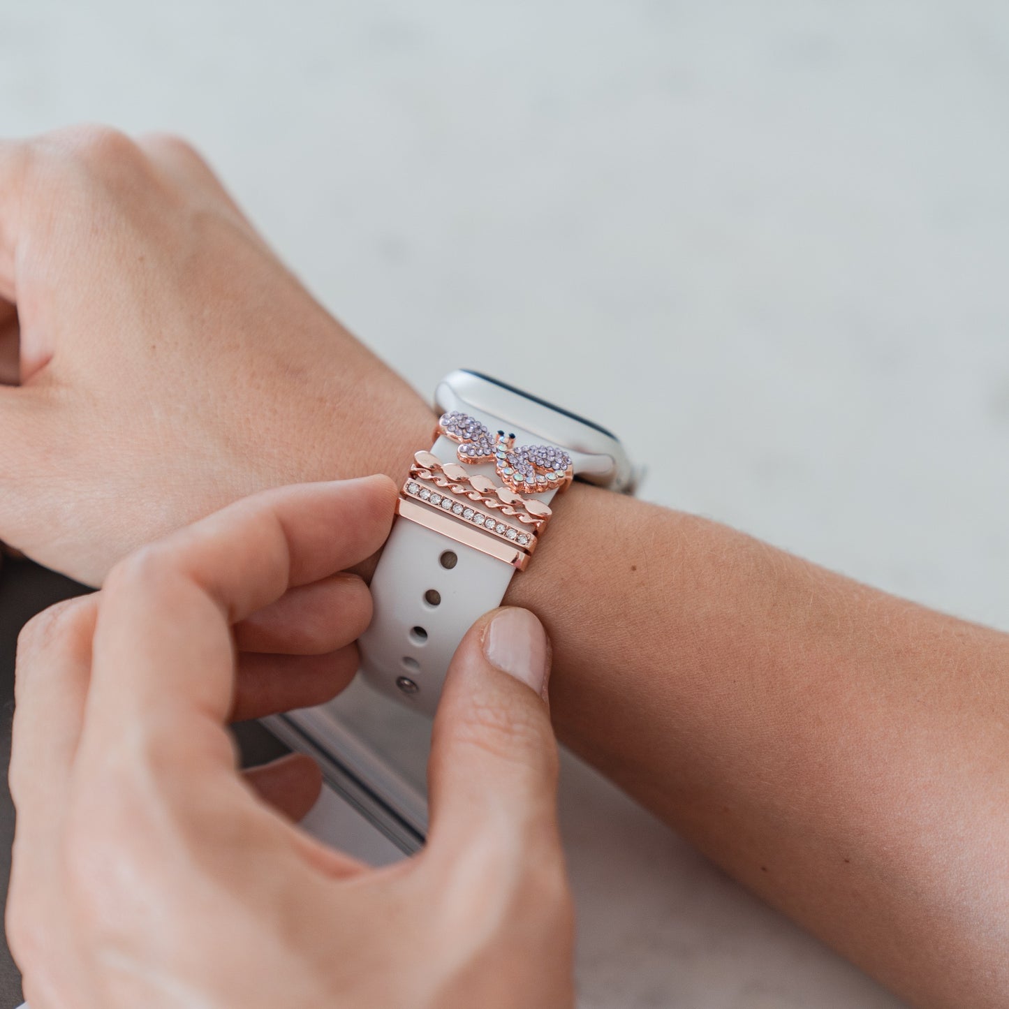 Eine Person passt ein weißes Apple Watch-Armband an, das mit Apple Watch-Armband-Charms von Arktisband verziert ist und übereinander gestapelte Schmetterlings- und Kronendesigns in Roségold von Arktisband aufweist, um vor einem hellen Hintergrund einen individuellen Look zu erzielen.