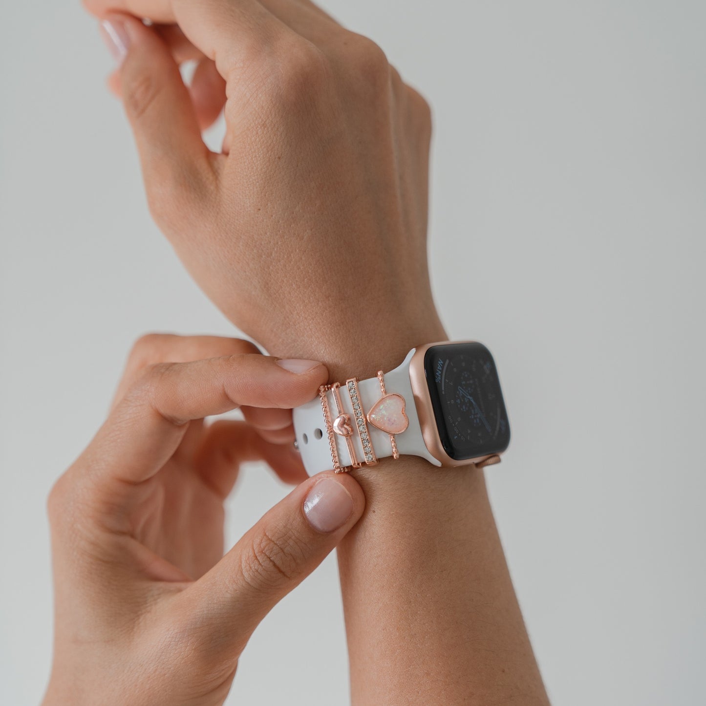 Jemand befestigt vor einem schlichten hellen Hintergrund eine weiße Arktisband-Smartwatch mit schwarzem Bildschirm, die mit mehreren rosa und goldenen Arktisband-Apple-Watch-Armband-Charms verziert ist, an seinem Handgelenk.