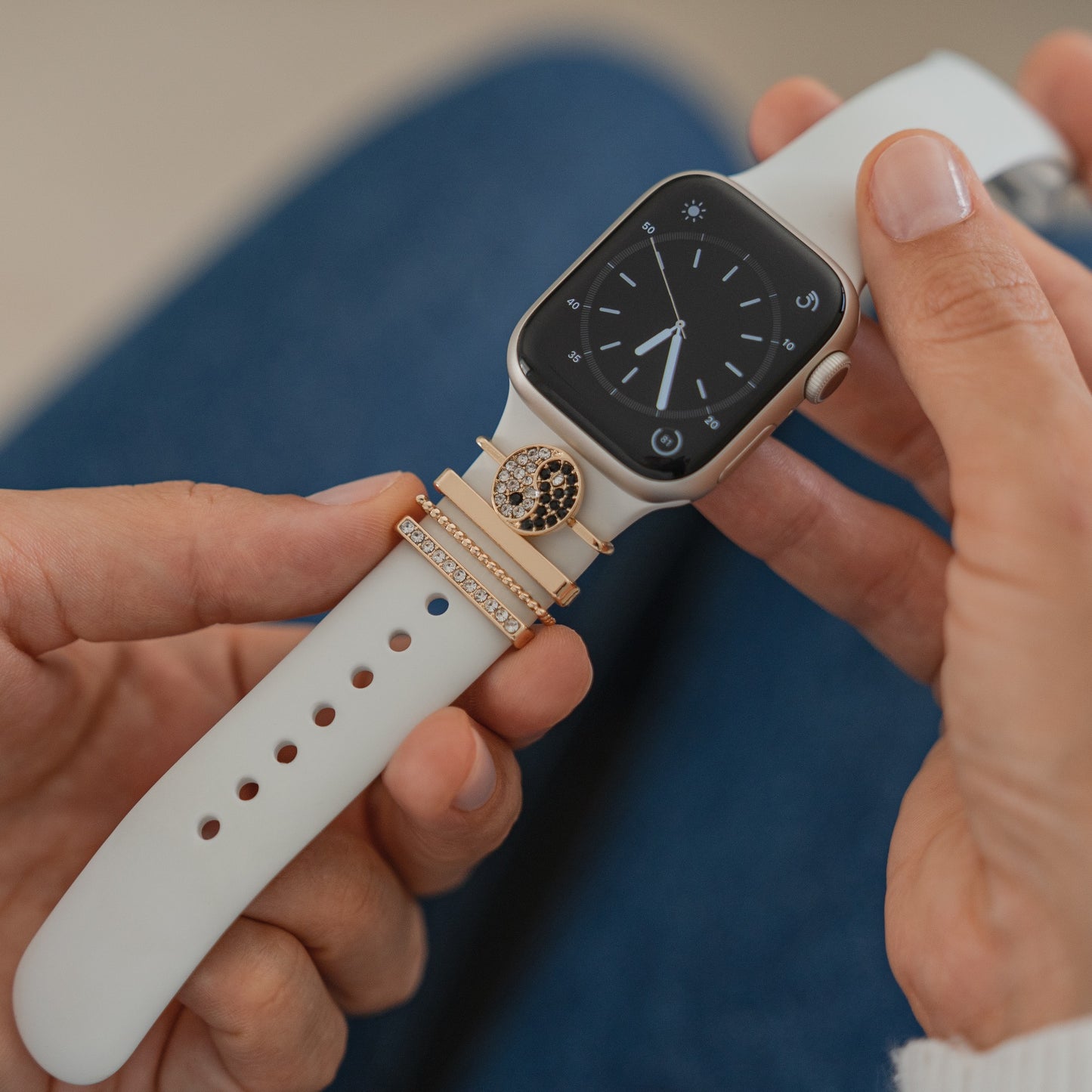 Eine Person passt ein weißes Apple Watch-Armband mit einem schwarzen Bildschirm an, auf dem eine analoge Uhr und drei arktisband Apple Watch-Armband-Charms von arktisband zu sehen sind, darunter einer, der mit kleinen Kristallen verziert ist.