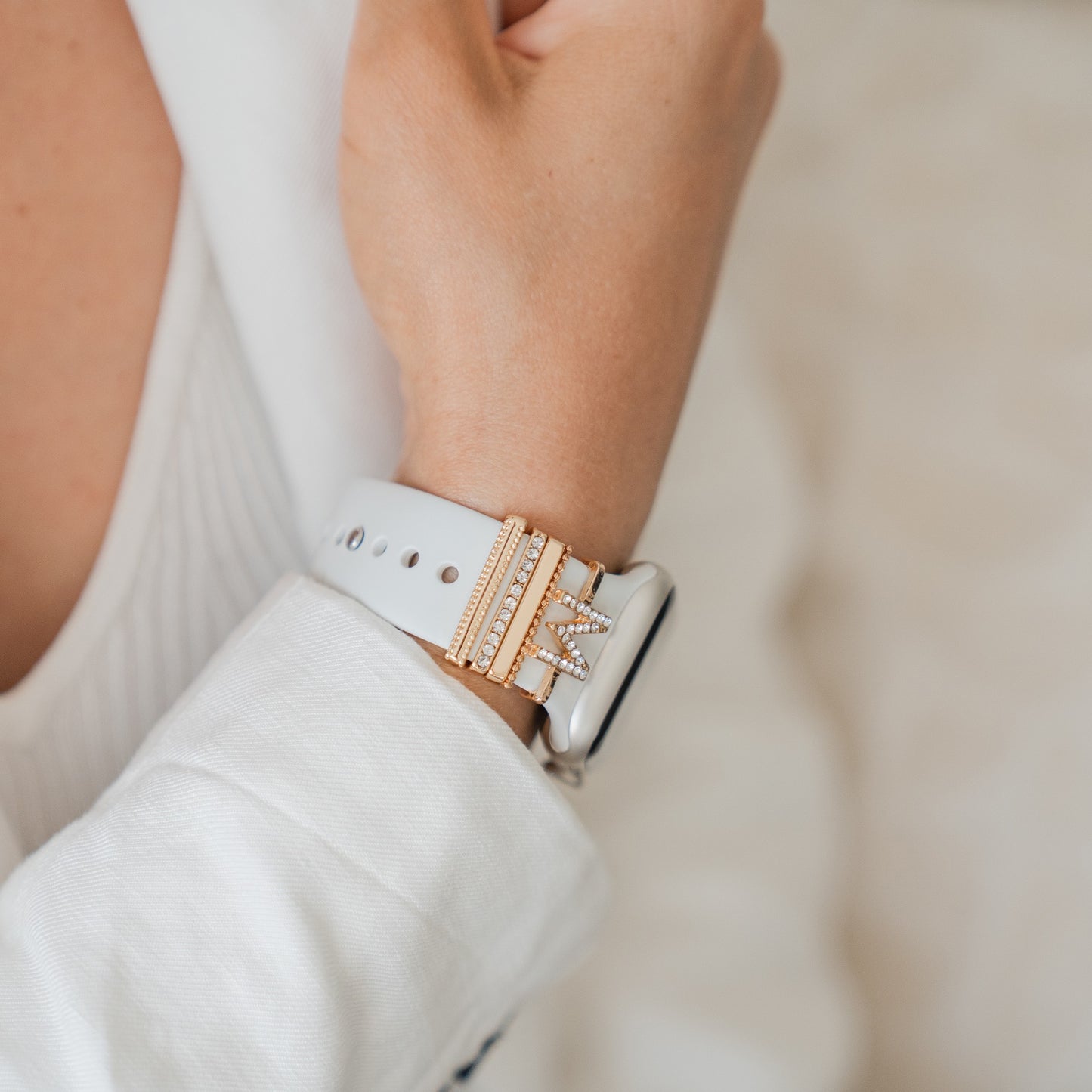 Eine Person trägt ein weißes Langarmshirt und eine weiße Smartwatch mit Arktisband Apple Watch Armband Charms von Arktisband sowie Gold- und Diamantbändern am Armband. Der Hintergrund ist unscharf.