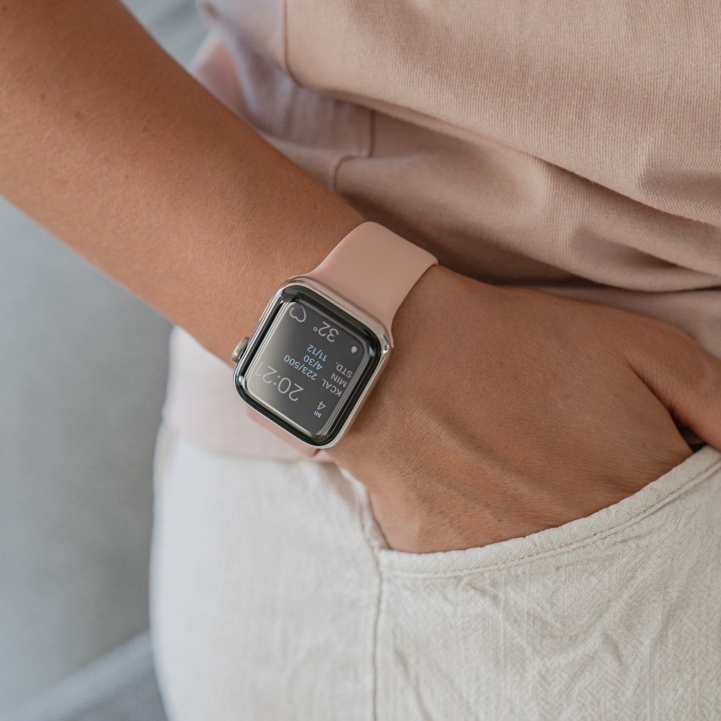 Eine Person, die ein Arktisband Apple Watch Silikonarmband „Aqua“ trägt, steht mit der Hand in der Tasche einer cremefarbenen Hose und auf dem Bildschirm der Apple Watch werden Trainingsstatistiken angezeigt.