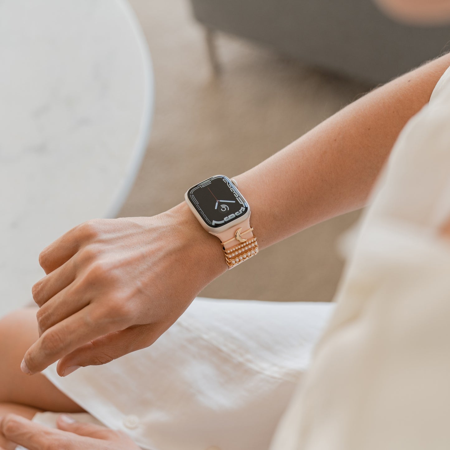 Eine Person mit einem arktisband Apple Watch Armband-Charm an einem hellen Armband trägt es am linken Handgelenk und zeigt ein Musik-App-Symbol. Sie sitzt in heller Kleidung an einem weißen runden Tisch und genießt dieses individuell gestaltbare arktisband Accessoire.