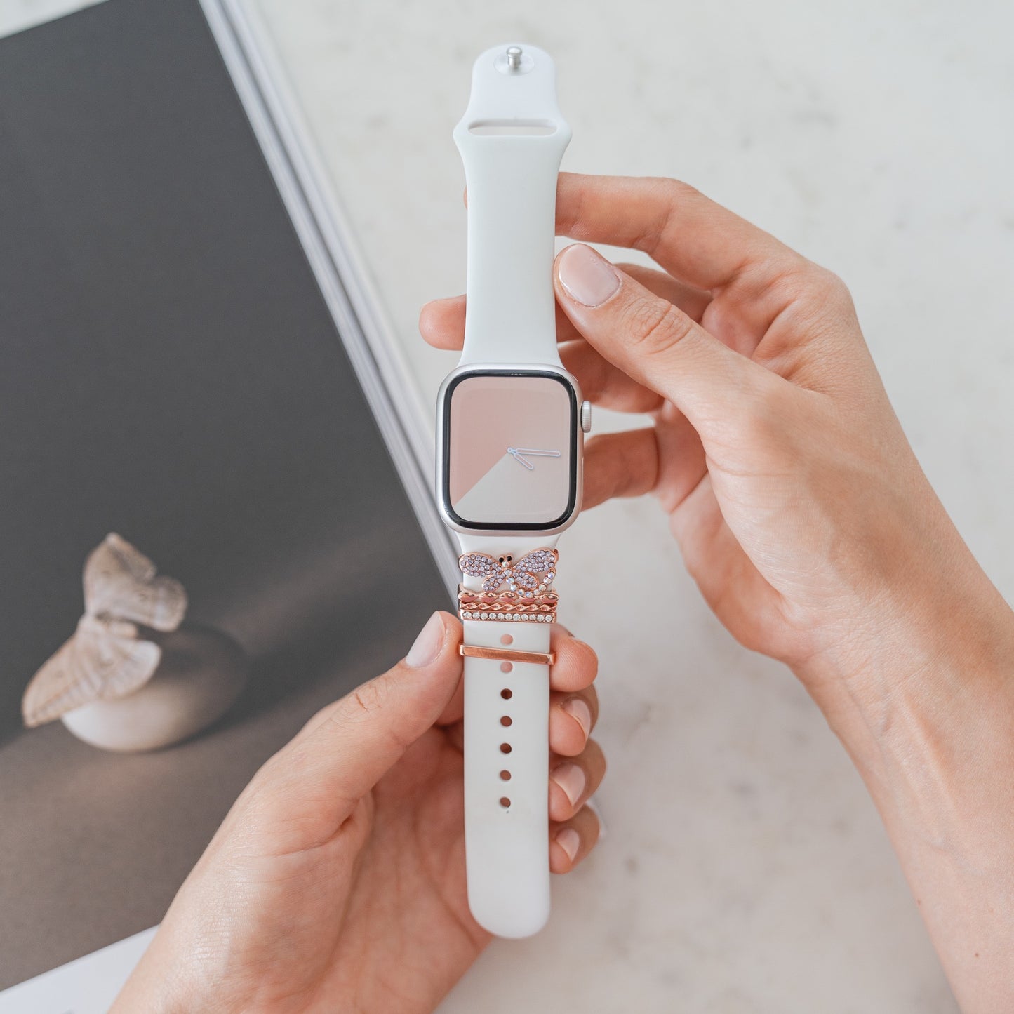 Jemand hält einen Arktisband Apple Watch Armband-Anhänger mit weißem Armband und rosa, diamantenbesetztem Akzent. Im Hintergrund liegt ein Arktisband-Buch mit Schmetterlingsmotiv auf einer glatten weißen Marmoroberfläche.