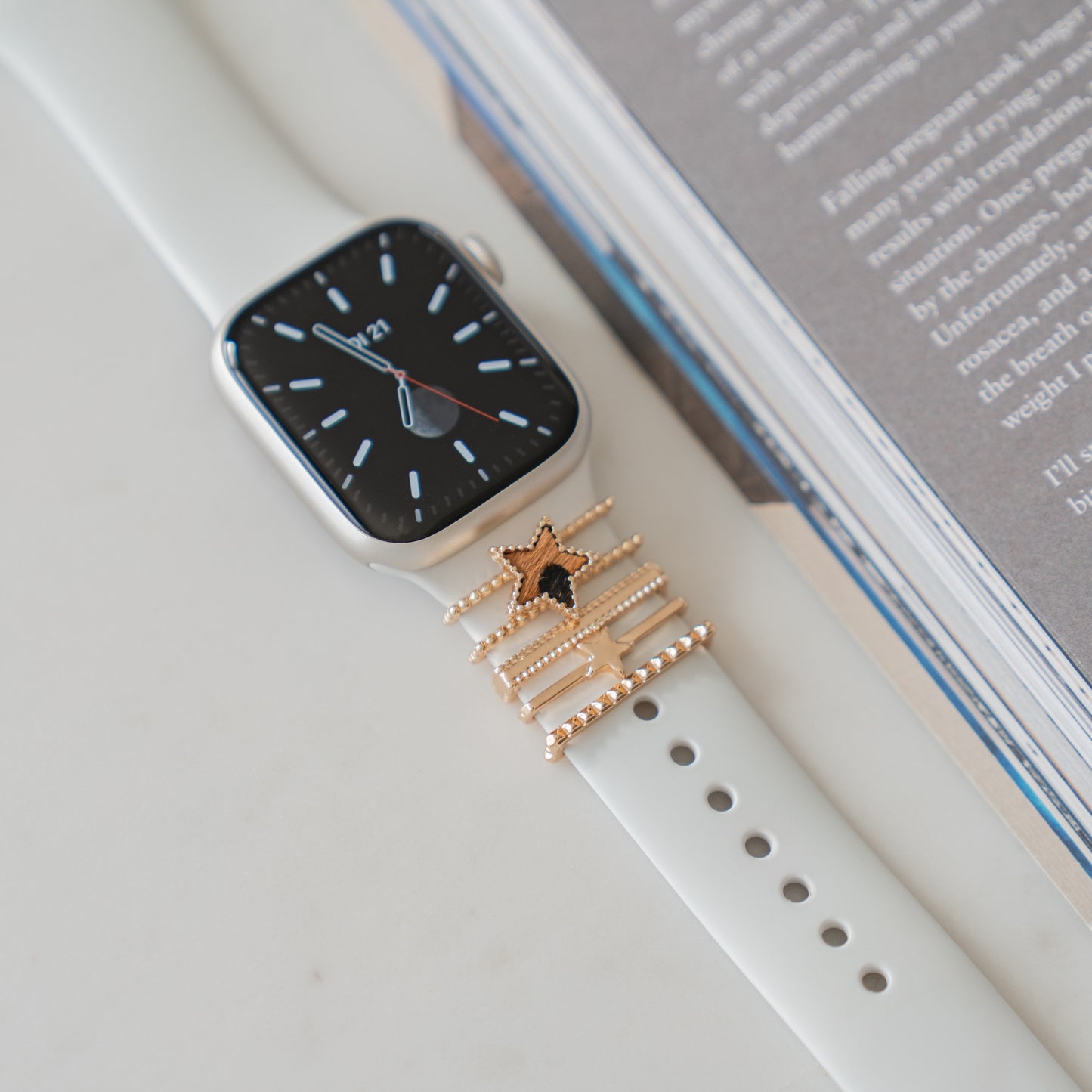 Eine weiße Apple Watch mit schwarzem Display liegt neben einem offenen Buch. Das Armband mit goldenem Sternanhänger unterstreicht, wie sich mit den arktisband Apple Watch Armbandanhängern der Look individuell gestalten lässt.