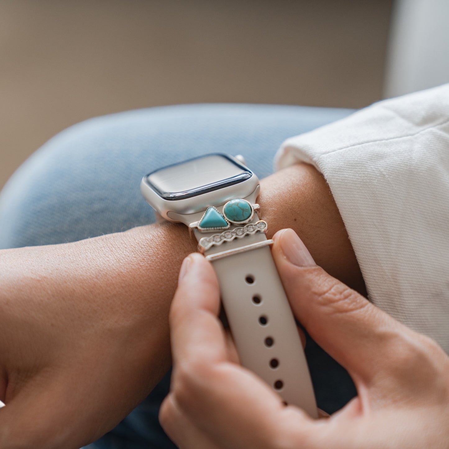 Jemand rückt sein Arktisband Apple Watch Armband Charms-Armband von Arktisband zurecht, das aus einem beigen Armband mit türkisfarbenen Dreiecken und runden Steinanhängern besteht, während er helle Kleidung trägt.