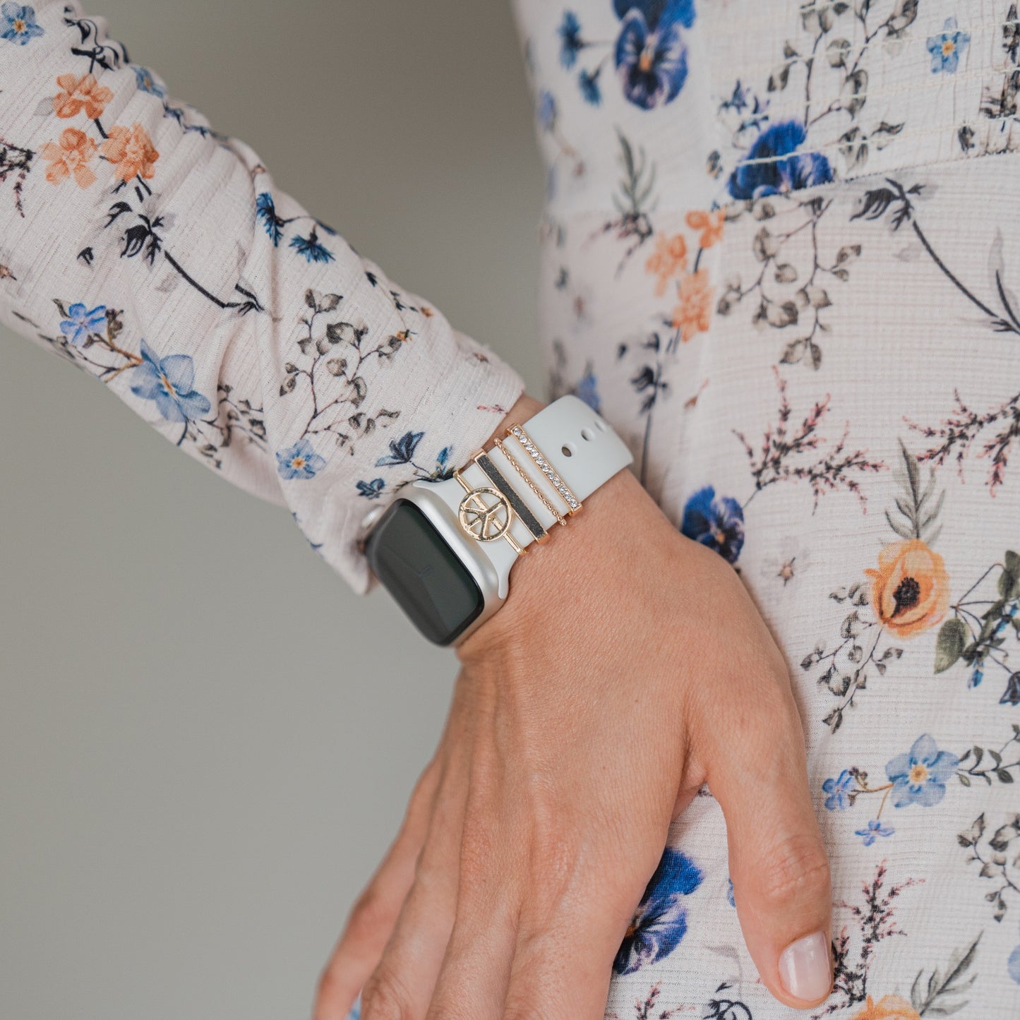 Eine Person in einem langärmeligen Blumenkleid stützt ihre Hand auf die Hüfte und trägt eine Apple Watch, die mit arktisband Apple Watch Armband Charms von arktisband verziert ist, gepaart mit goldenen und weißen Armbändern.