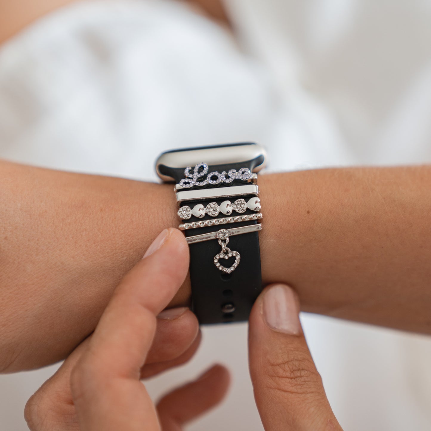 Eine Nahaufnahme einer Person, die ihre Arktisband-Apple-Watch-Armband-Charms von Arktisband an einem schwarzen Smartwatch-Armband anpasst, das mit silbernen und Strass-Charms wie „Love“, Herzen und einem baumelnden Herz-Charm versehen ist.