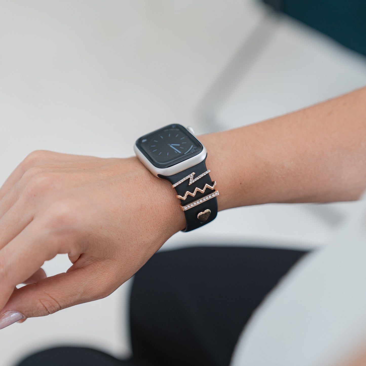 Eine Person trägt eine Smartwatch mit einem schwarzen Arktisband, verziert mit goldenen und strassbesetzten Zickzack- sowie herzförmigen Arktisband-Apple-Watch-Armbandanhängern. Der Bildschirm ist ausgeschaltet, die Hand ruht auf dem Schoß.