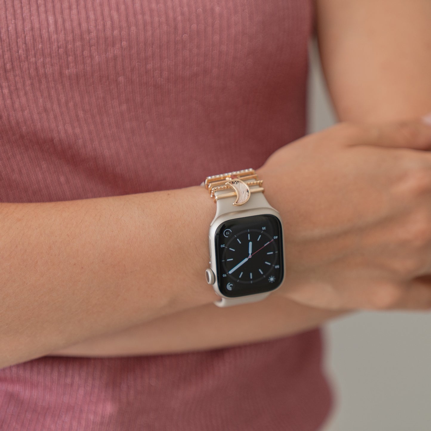 Eine Person, die ein rosa geripptes ärmelloses Top und ein Arktisband Apple Watch Armband Charms-Armband am linken Handgelenk trägt, steht mit verschränkten Armen da, während die Smartwatch die Uhrzeit anzeigt.