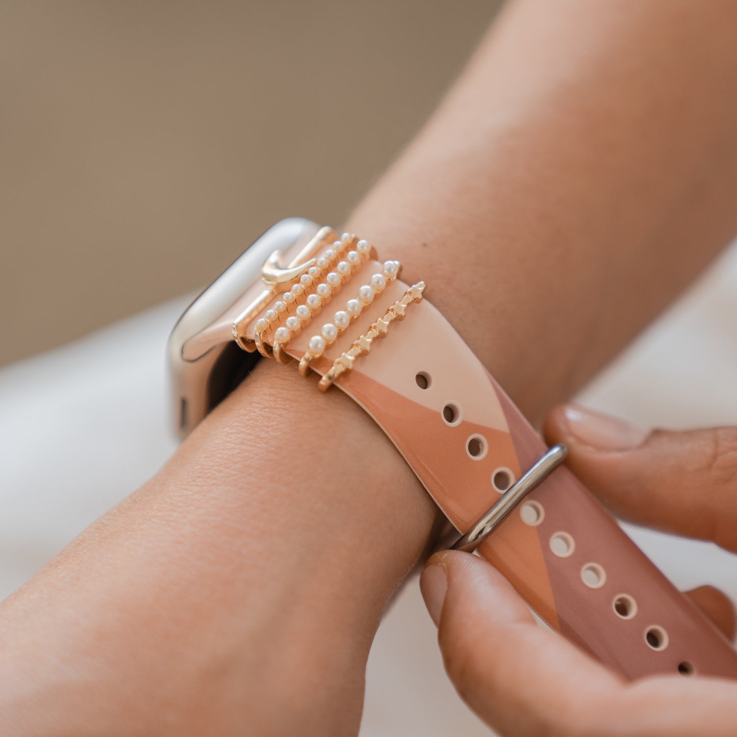 Eine Nahaufnahme eines Handgelenks mit einer individuell gestaltbaren Arktisband-Smartwatch mit rosa und braunen Armbändern, verziert mit Arktisband-Apple-Watch-Armband-Charms, die um einen Teil des Armbands gewickelt sind.