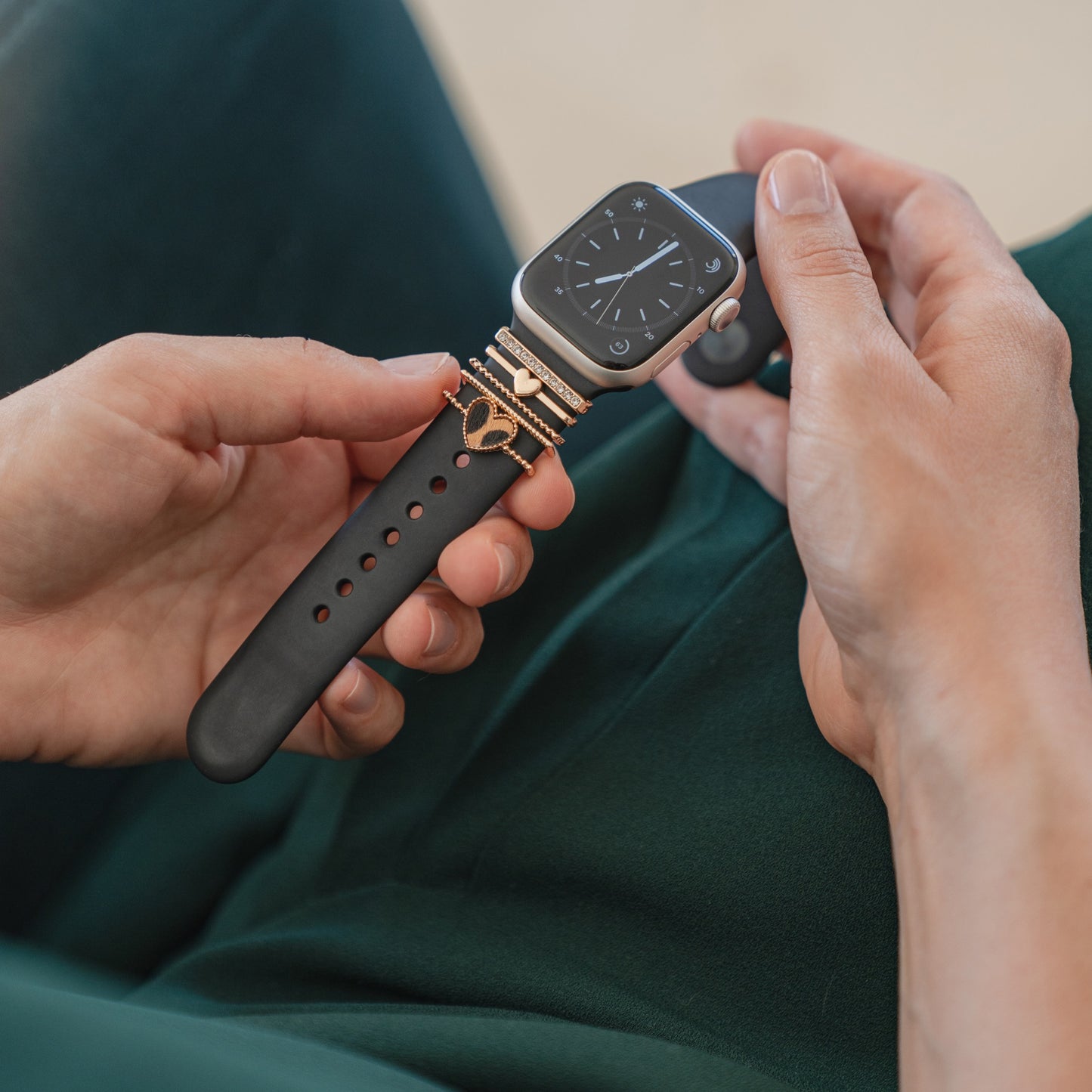 Eine Person hält eine schwarze Apple Watch mit rechteckigem Zifferblatt, verziert mit Arktisband Apple Watch Armband Charms von Arktisband, und sitzt auf dunkelgrünem Stoff.