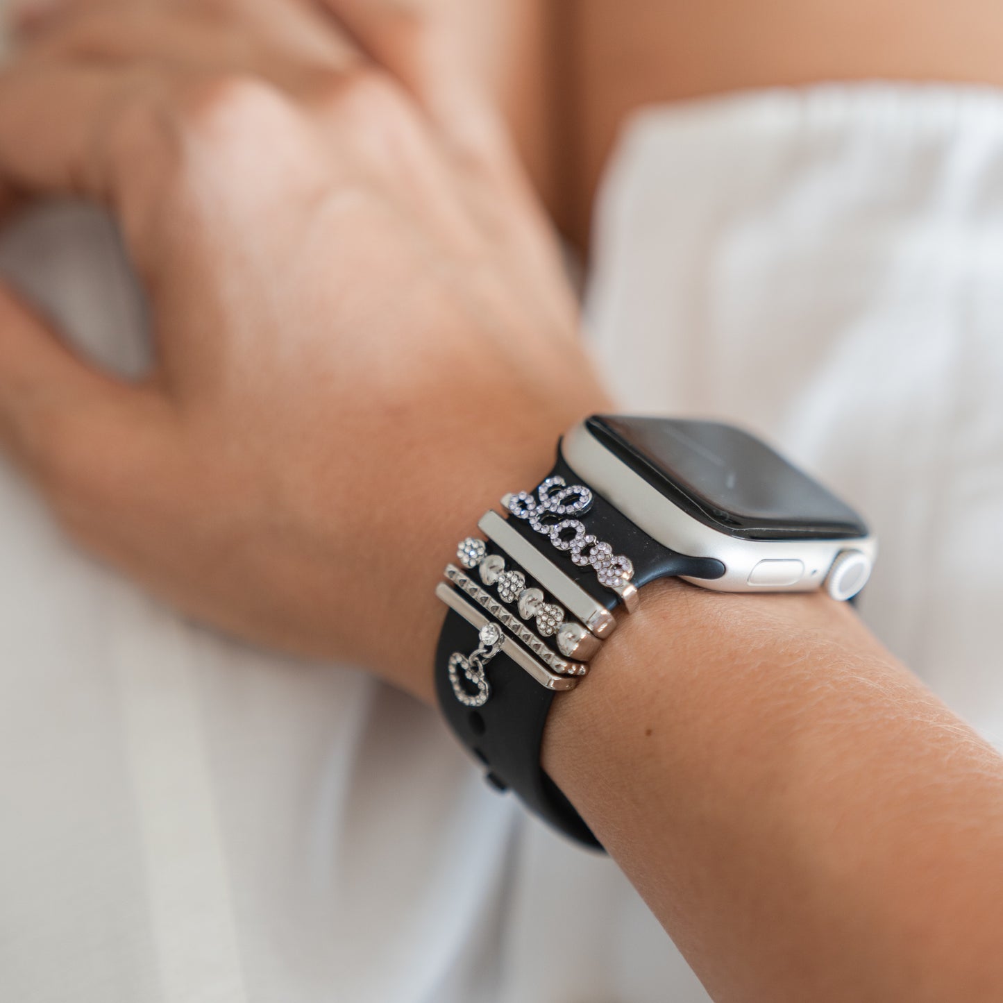 Nahaufnahme eines Handgelenks mit einem Arktisband Apple Watch Armband Charms von Arktisband, mit individuell anpassbaren Anhängern aus Silber und Strass, vor einem verschwommenen weißen Stoffhintergrund.