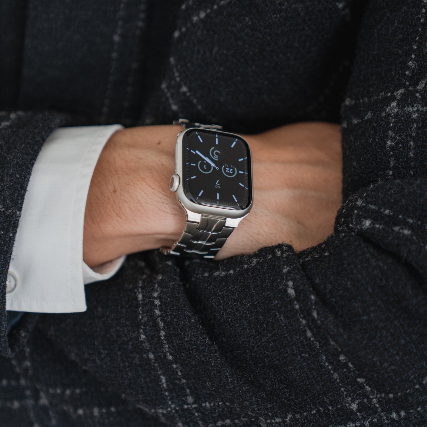 Eine Person in einem dunkelkarierten Blazer und weißen Hemdmanschetten trägt das arktisband Apple Watch Edelstahlarmband „Momento“. Die silberne Smartwatch von arktisband zeigt Uhrzeit und Informationen auf einem dunklen Display an.