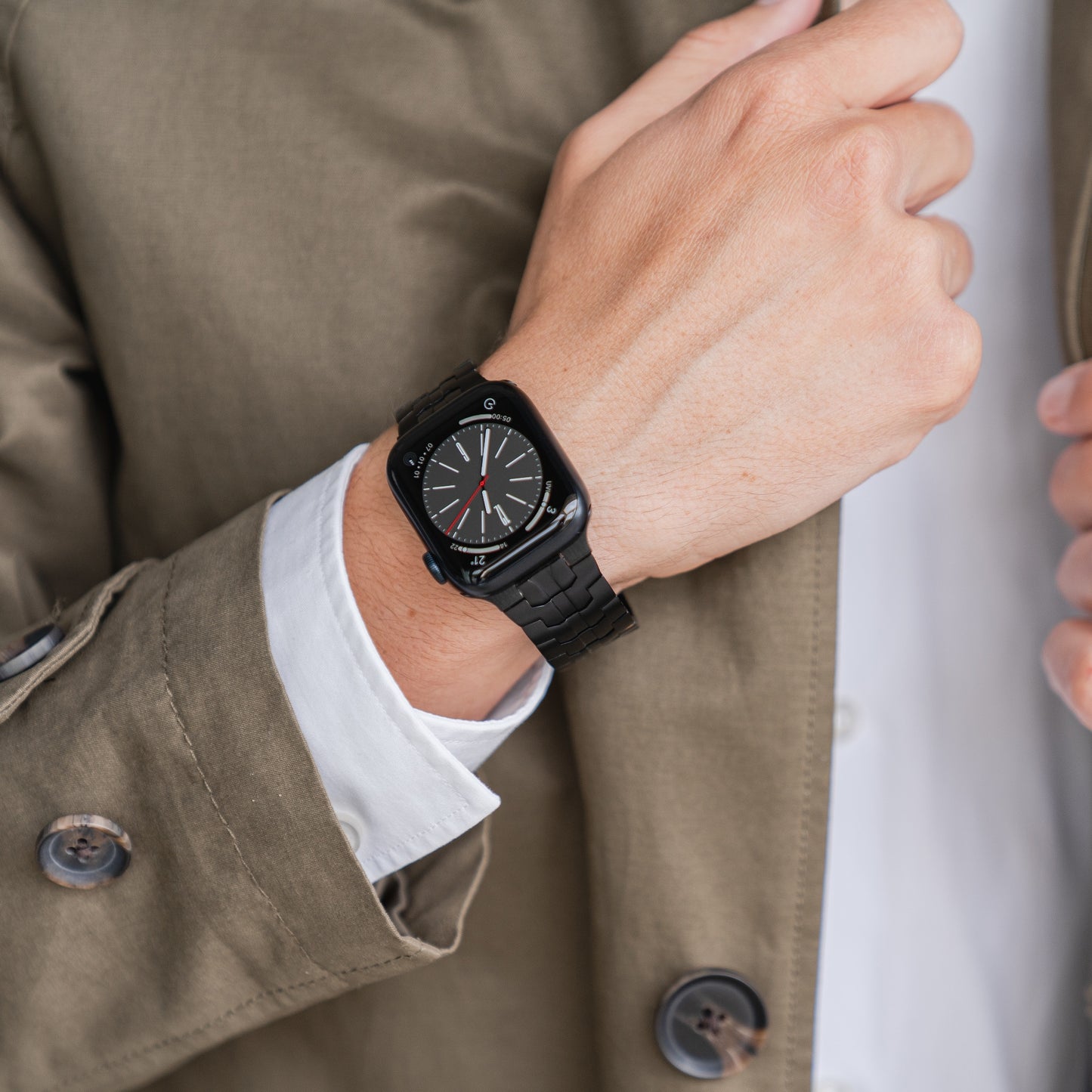 Eine Person in einem beigen Mantel und einem weißen Hemd ist von der Brust abwärts zu sehen. Sie trägt ein arktisband Apple Watch Edelstahlarmband „Momento“ von arktisband und eine schwarze Smartwatch im Analogstil am linken Handgelenk.
