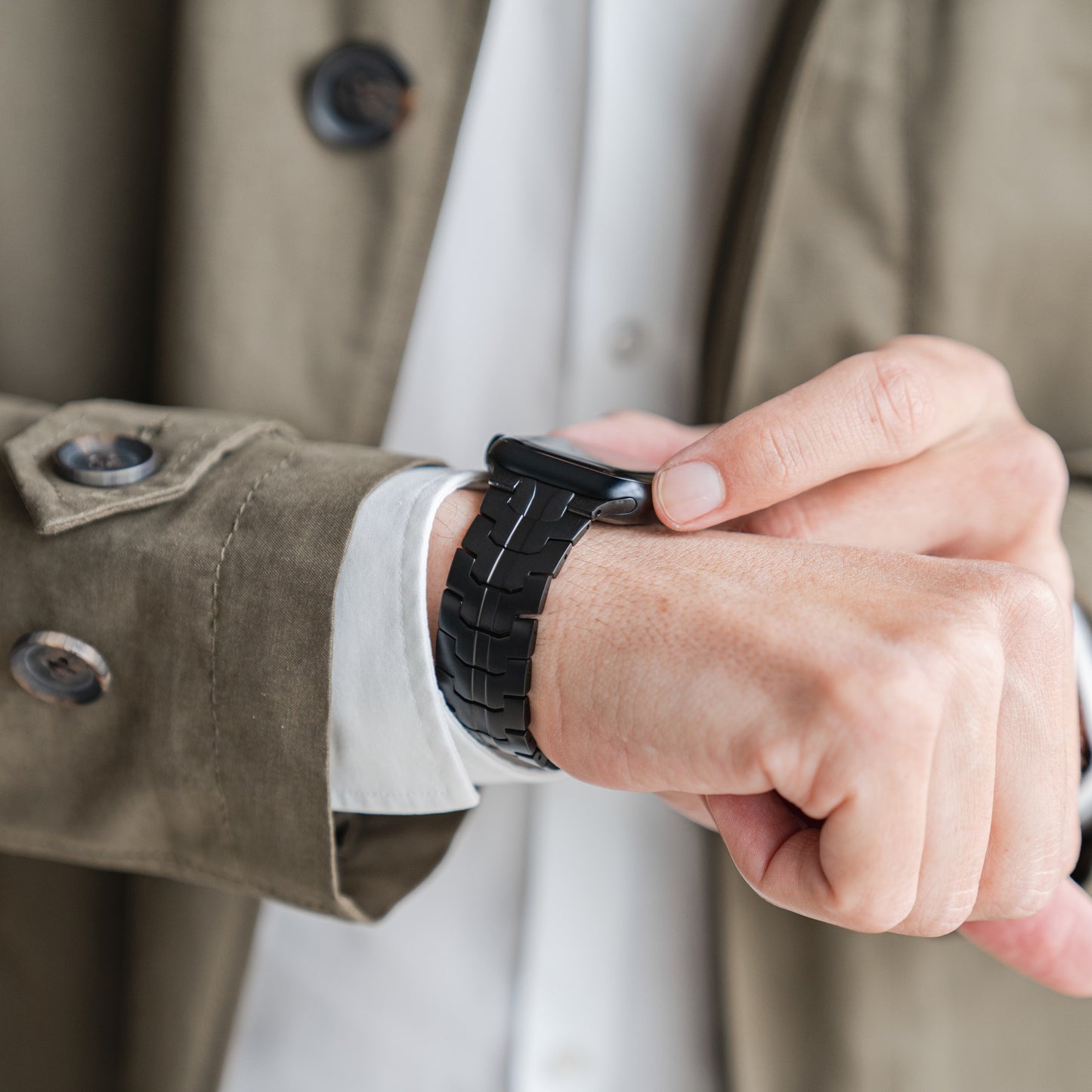 Eine Person in beigem Trenchcoat und weißem Hemd rückt das arktisband Apple Watch Edelstahlarmband „Momento“ an ihrem linken Handgelenk zurecht. Der Fokus liegt auf den Zeigern, der Uhr und den deutlich sichtbaren Mantelknöpfen.