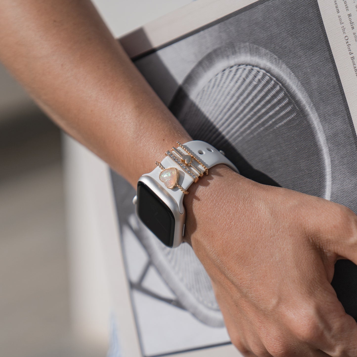 Eine Nahaufnahme eines Handgelenks mit Arktisband-Apple-Watch-Armbandanhängern mit herzförmigem Anhänger, goldenen Armbändern und einem Arktisband-Armband. Die Person hält eine Zeitung mit einem kreisförmigen Muster auf der Titelseite.