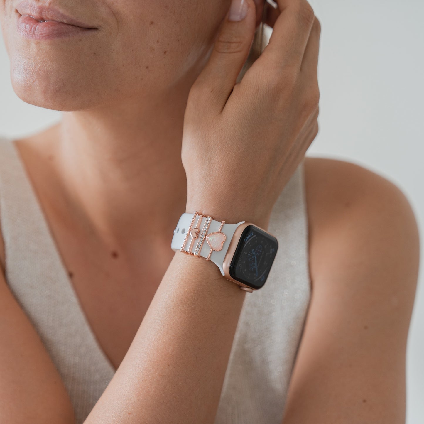 Eine Person in einem ärmellosen beigen Oberteil legt ihre Hand auf ihr Gesicht und präsentiert eine Arktisband-Smartwatch mit Akzenten in Roségold und Arktisband-Apple-Watch-Armbandanhängern an ihrem Handgelenk.