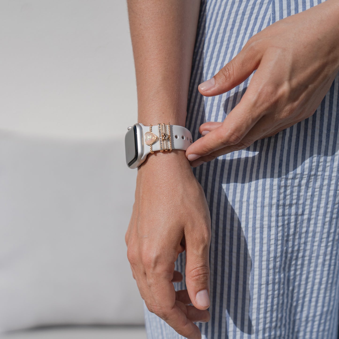 Eine Person in einem blau-weiß gestreiften Kleid rückt ihre Arktisband Apple Watch Armband Charms von Arktisband zurecht, die sie mit goldenen und perlenbesetzten Armbändern kombiniert. Der sanft beleuchtete, neutrale Hintergrund unterstreicht die Szene.