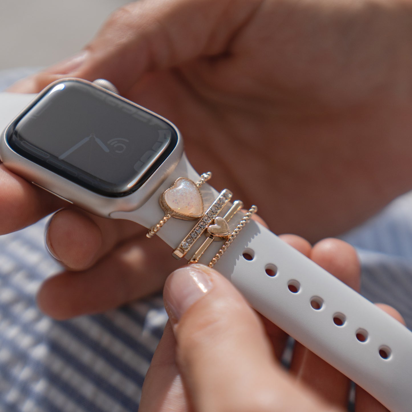 Eine Person hält eine Apple Watch mit weißem Arktisband, verziert mit Arktisband Apple Watch Armband Charms – Gold, Perlen und herzförmigen Perlen. Das Zifferblatt ist schwarz und scheint sich im Standby-Modus zu befinden.