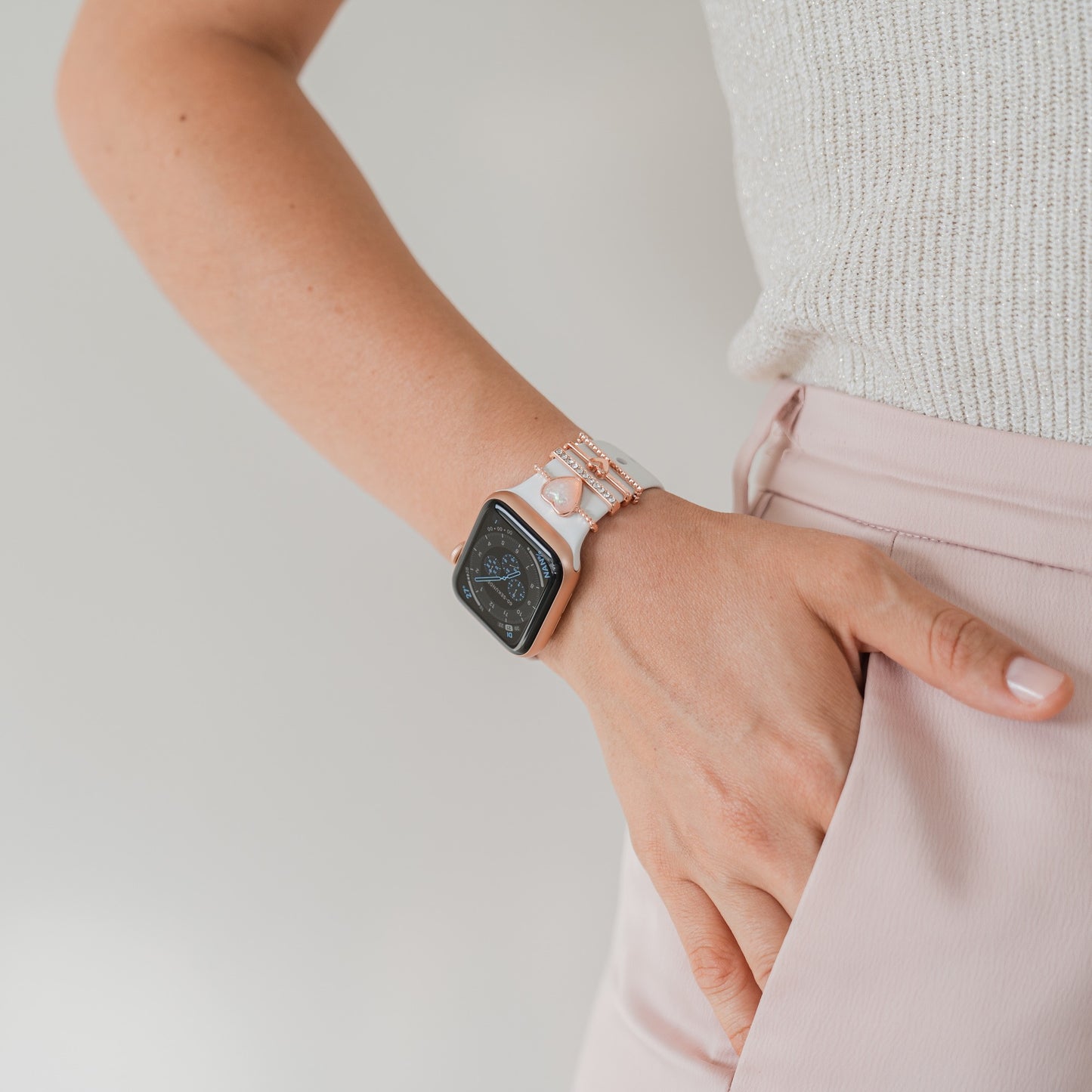 Eine Person in einem hellen ärmellosen Top und einer rosa Hose, die rechte Hand in der Tasche, trägt ein Arktisband Apple Watch Armband Charms und eine Smartwatch am Handgelenk. Der Hintergrund ist schlicht und hell.