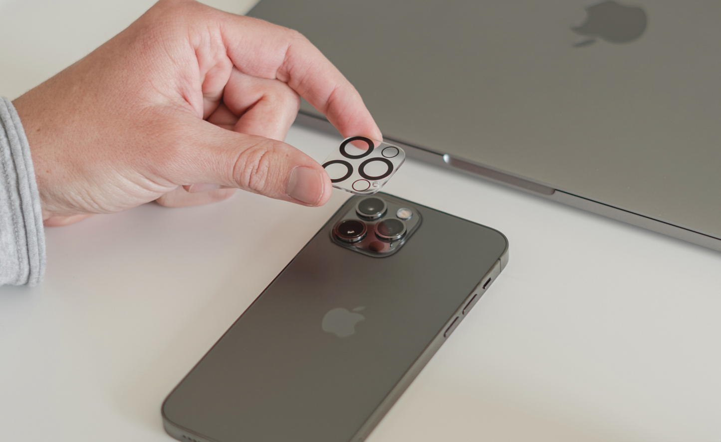 Eine Hand bringt einen ArktisPRO iPhone 13 Pro Kamera Protector von ArktisPRO auf der Rückseite eines schwarzen iPhones in der Nähe eines Laptops an und sorgt so für optimalen Kratzschutz.