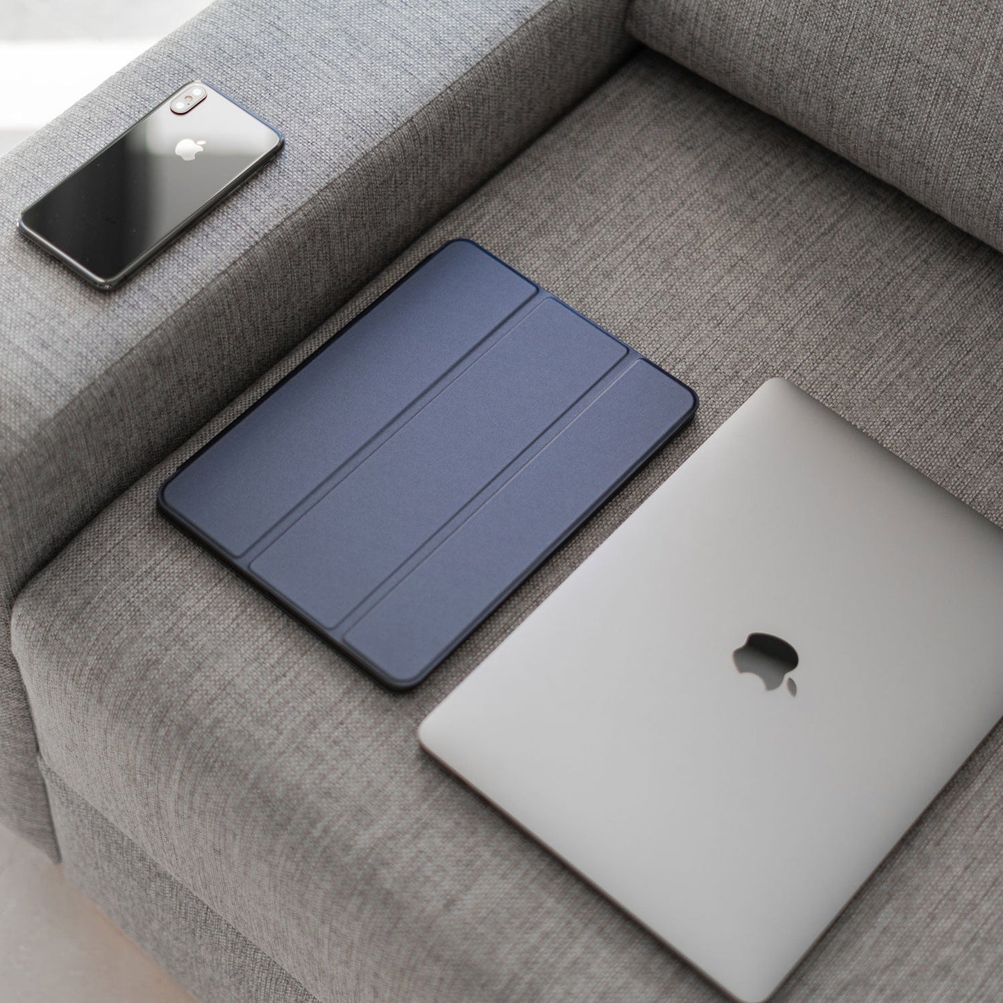 Ein schwarzes Smartphone, ein iPad 11" (2022/2025) im iCEO magnetischen Smart Case und ein geschlossenes silbernes Laptop liegen auf Armlehne und Sitz eines grauen Stoffsofas.