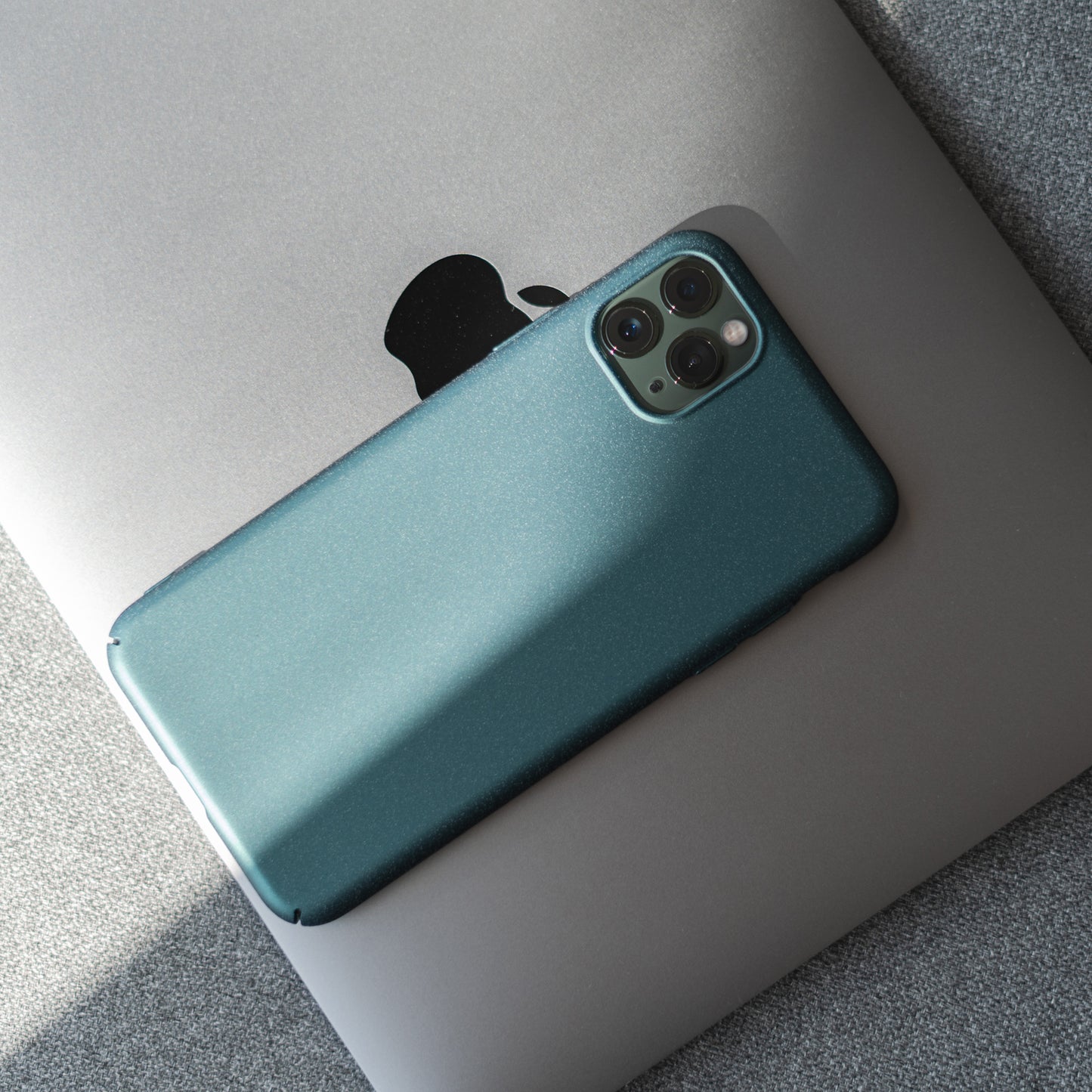 Ein iPhone 11 Pro mit dem ArktisPRO iPhone 11 Pro Hülle UltraSlim Hardcase in Grün ruht auf einem geschlossenen silbernen Laptop mit einem teilweisen Apple-Logo und bietet eleganten Schutz und unterstreicht gleichzeitig die Eleganz des Geräts.