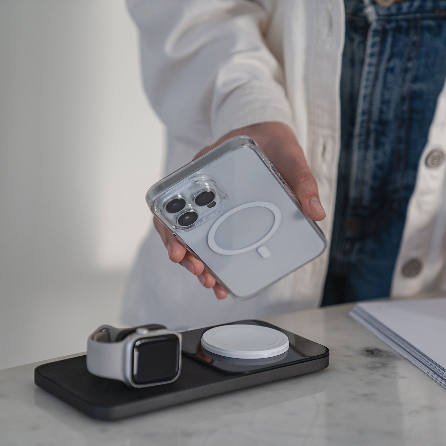 Eine Person in einem weißen Hemd hält ein MR PROTECT iPhone 14 Pro CLEAR Case mit MagSafe über eine kabellose Ladestation mit einer Smartwatch, alles auf einer Marmoroberfläche.