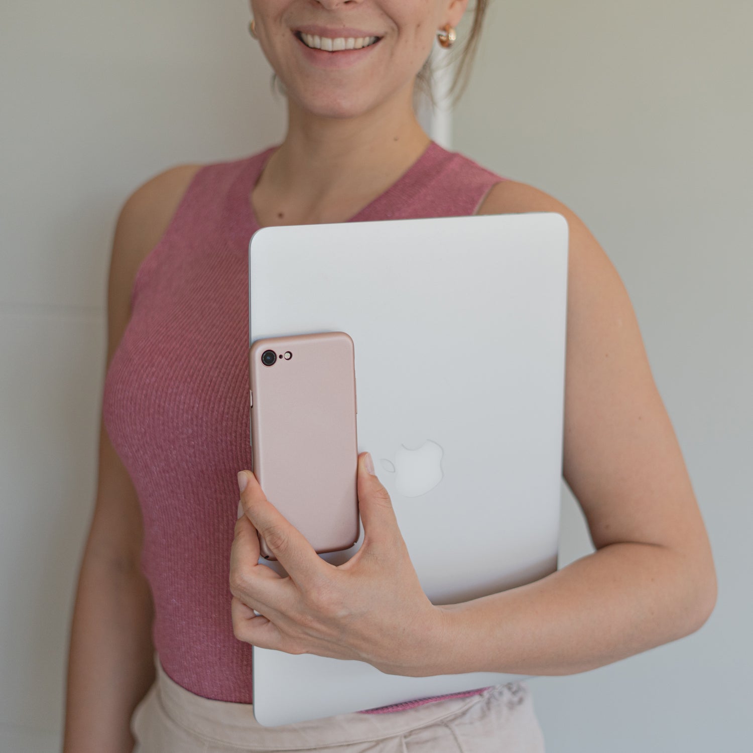 Eine Frau in einem ärmellosen rosa Top lächelt, während sie einen silbernen Laptop und ein rosa Smartphone hält, die durch das ArktisPRO iPhone 8 UltraSlim Hardcase von ArktisPRO geschützt sind.
