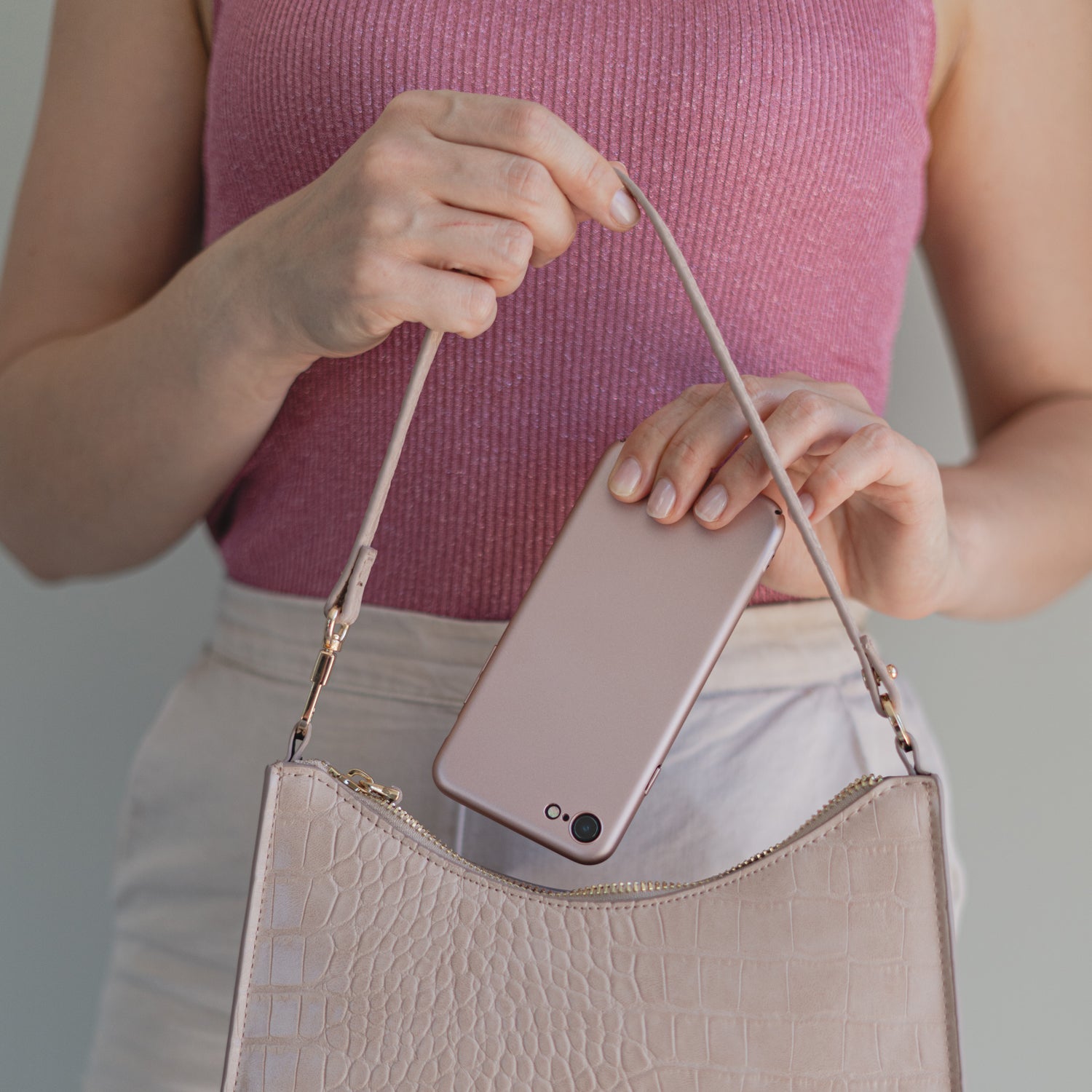 Eine Frau in einem rosa Oberteil und einem hellen Rock steckt ein rosa Smartphone mit einem ArktisPRO iPhone 8 UltraSlim Hardcase in eine beige Handtasche.