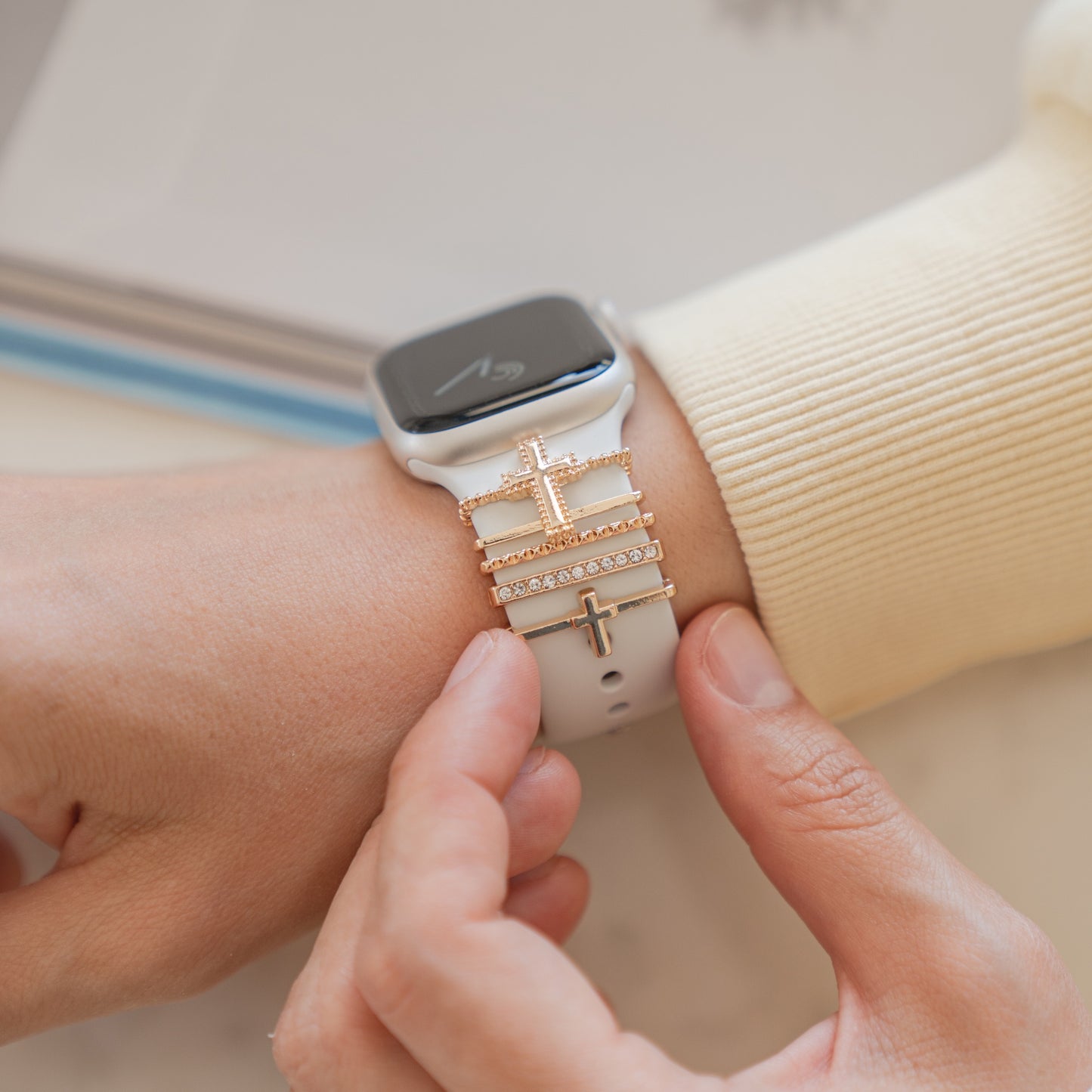 Eine Person trägt Arktisband Apple Watch Armband-Anhänger mit gestapelten goldenen Kreuzanhängern und Strasssteinen am Handgelenk und passt dabei individuell gestaltbare Anhänger am Arktisband-Armband an. Im Hintergrund ist ein unscharfes Notizbuch zu sehen.