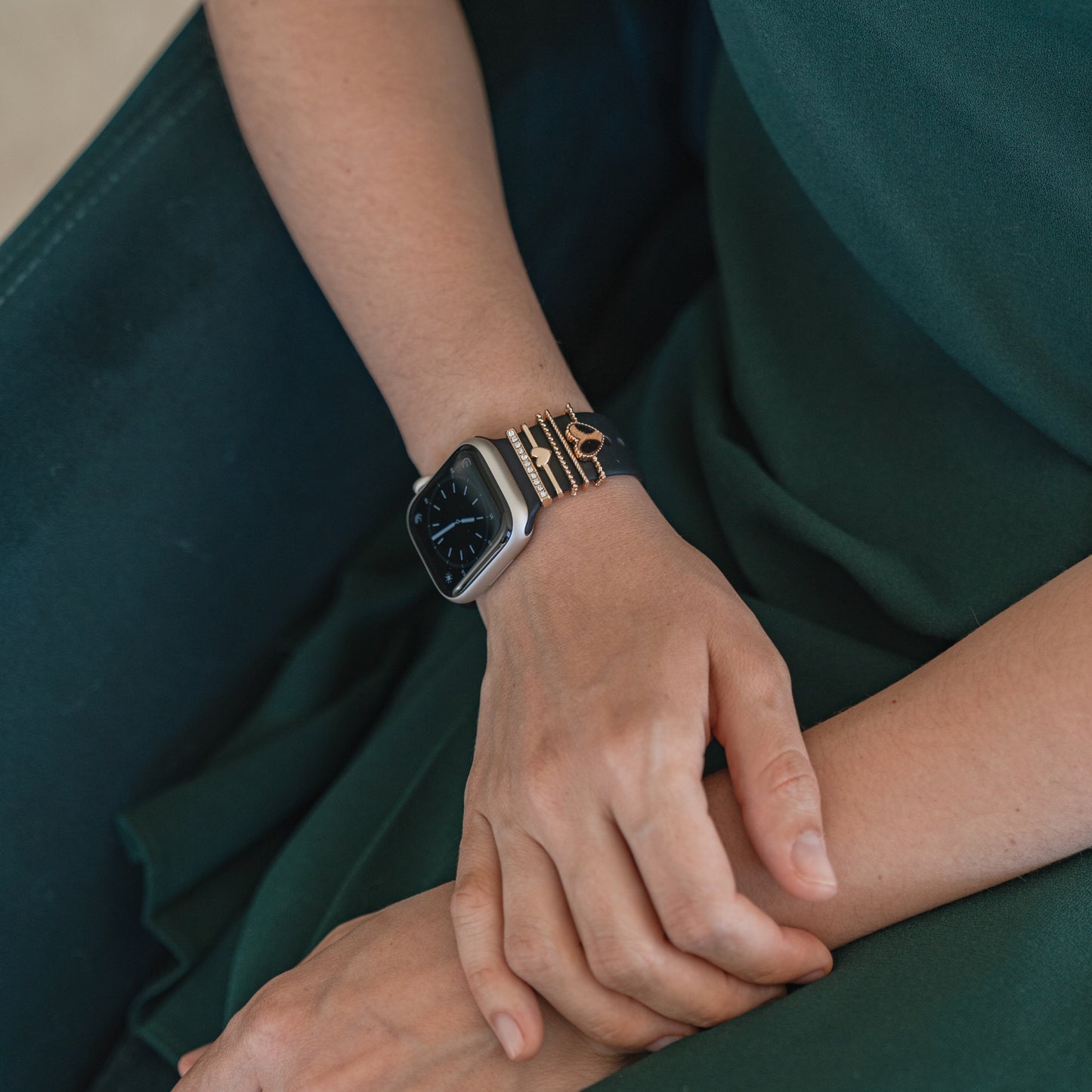 Eine Person in einem grünen Kleid sitzt mit den Händen im Schoß und zeigt eine Apple Watch mit Arktisband Apple Watch Armband Charms und goldenen Armbändern an ihrem linken Handgelenk.